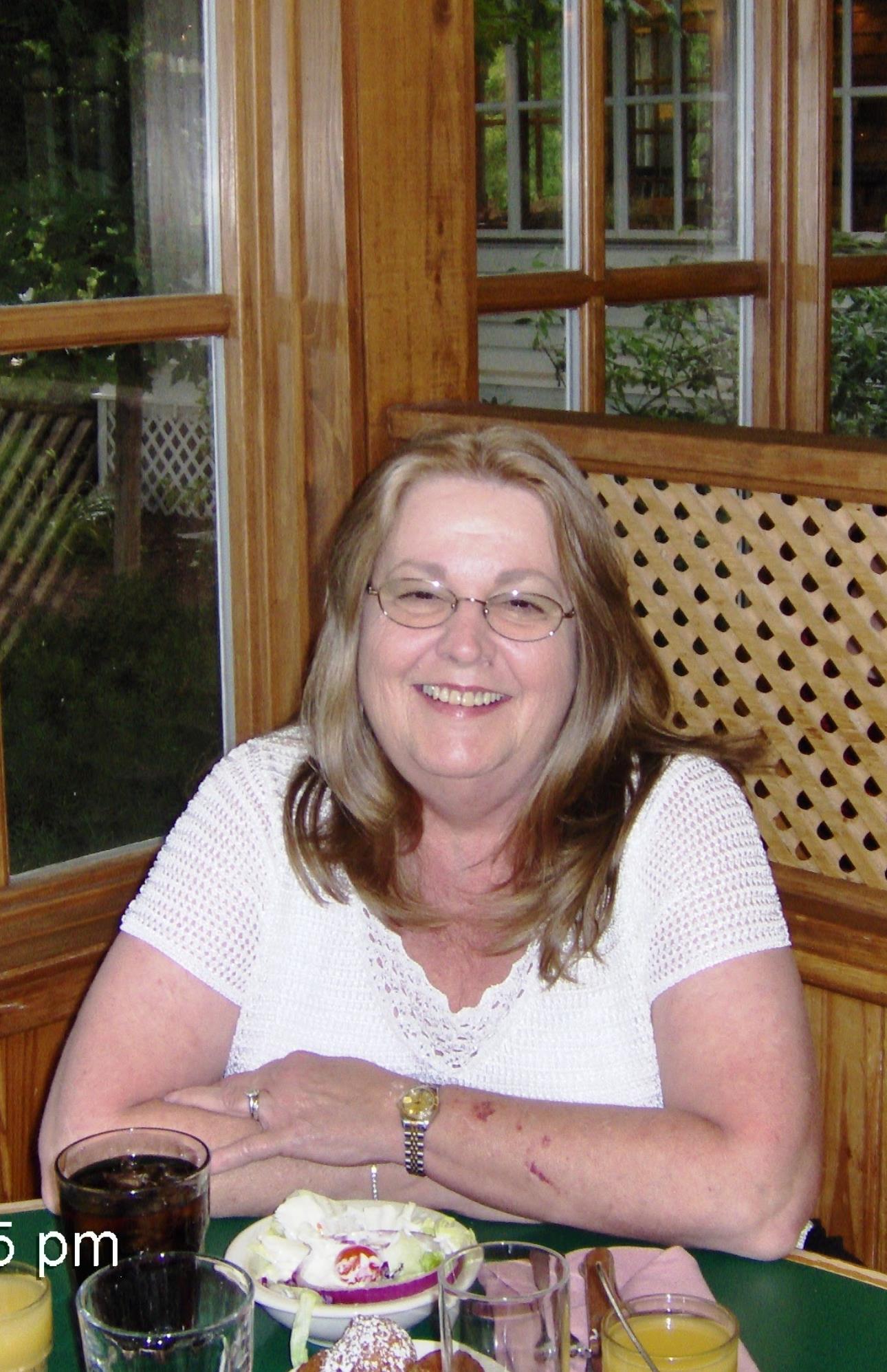 Woman with glasses smiles warmly while seated at a wooden table enjoying a meal.