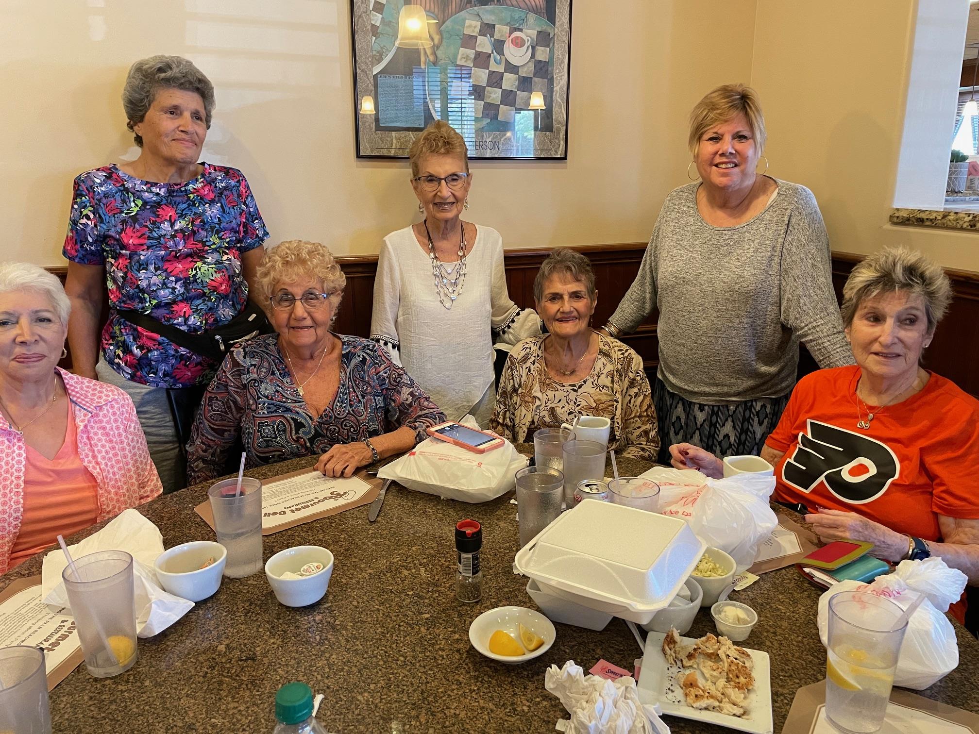 Seven women gather around a table, sharing food and conversation in a cozy restaurant atmosphere.