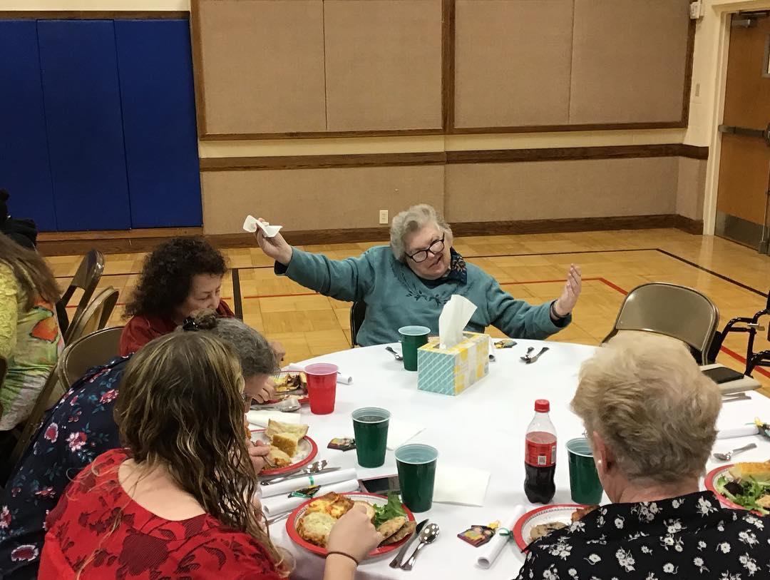 A group of elderly women share laughter and food at a community gathering in a lively atmosphere.