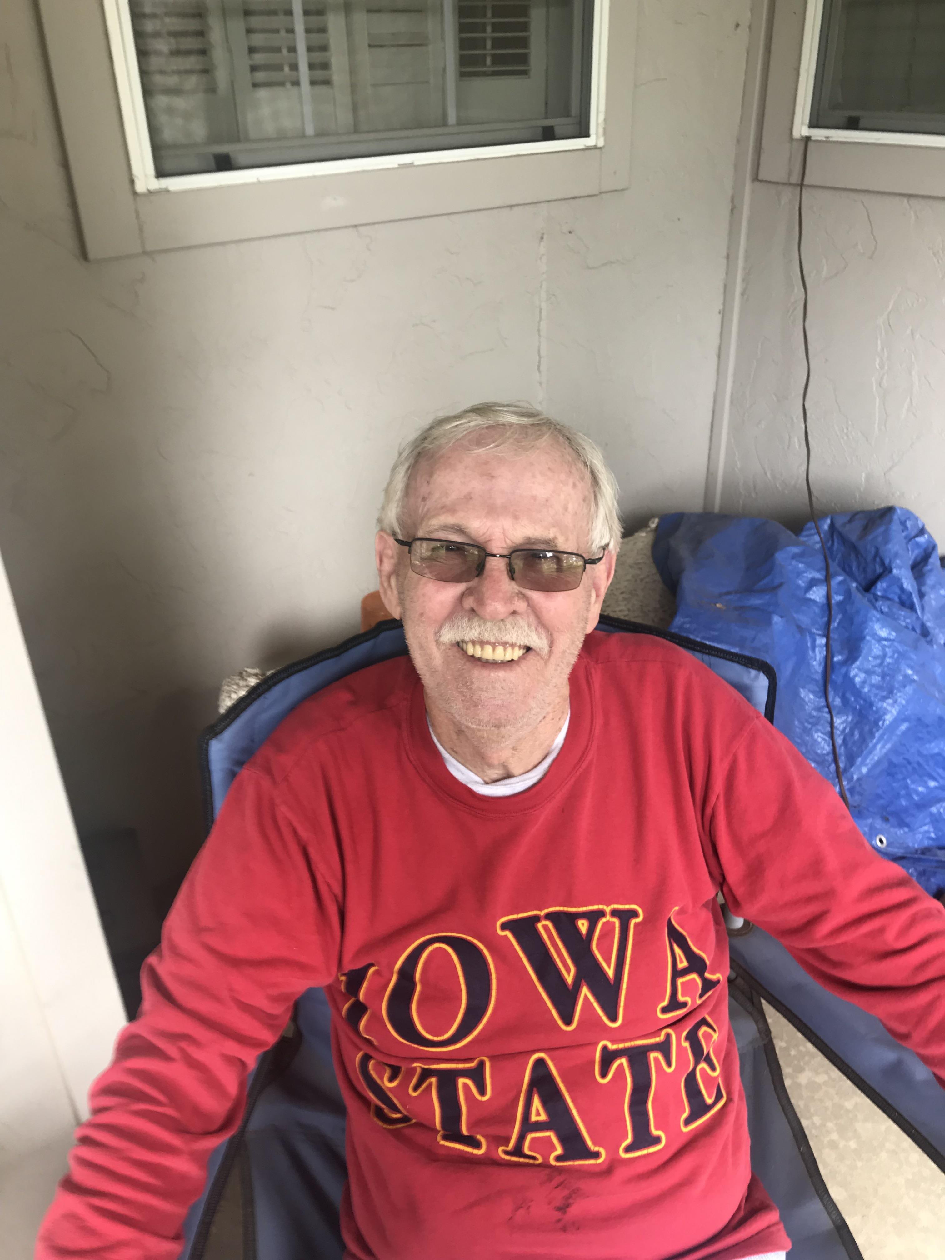 Elderly man sits comfortably in a chair, smiling joyfully while wearing a sports sweatshirt.