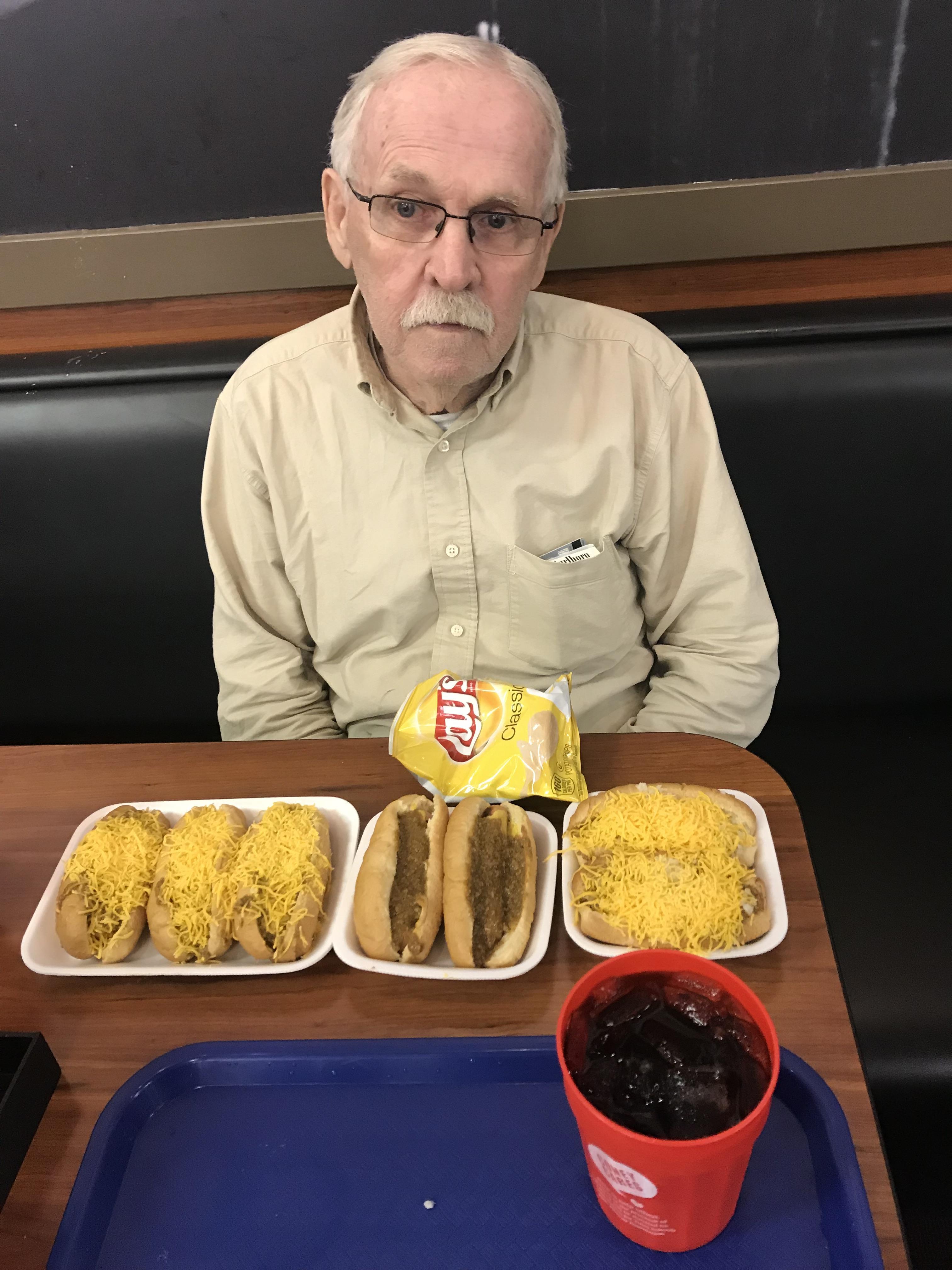 Man sits at a table with three hot dogs topped with cheese, chips, and a soda in a diner.