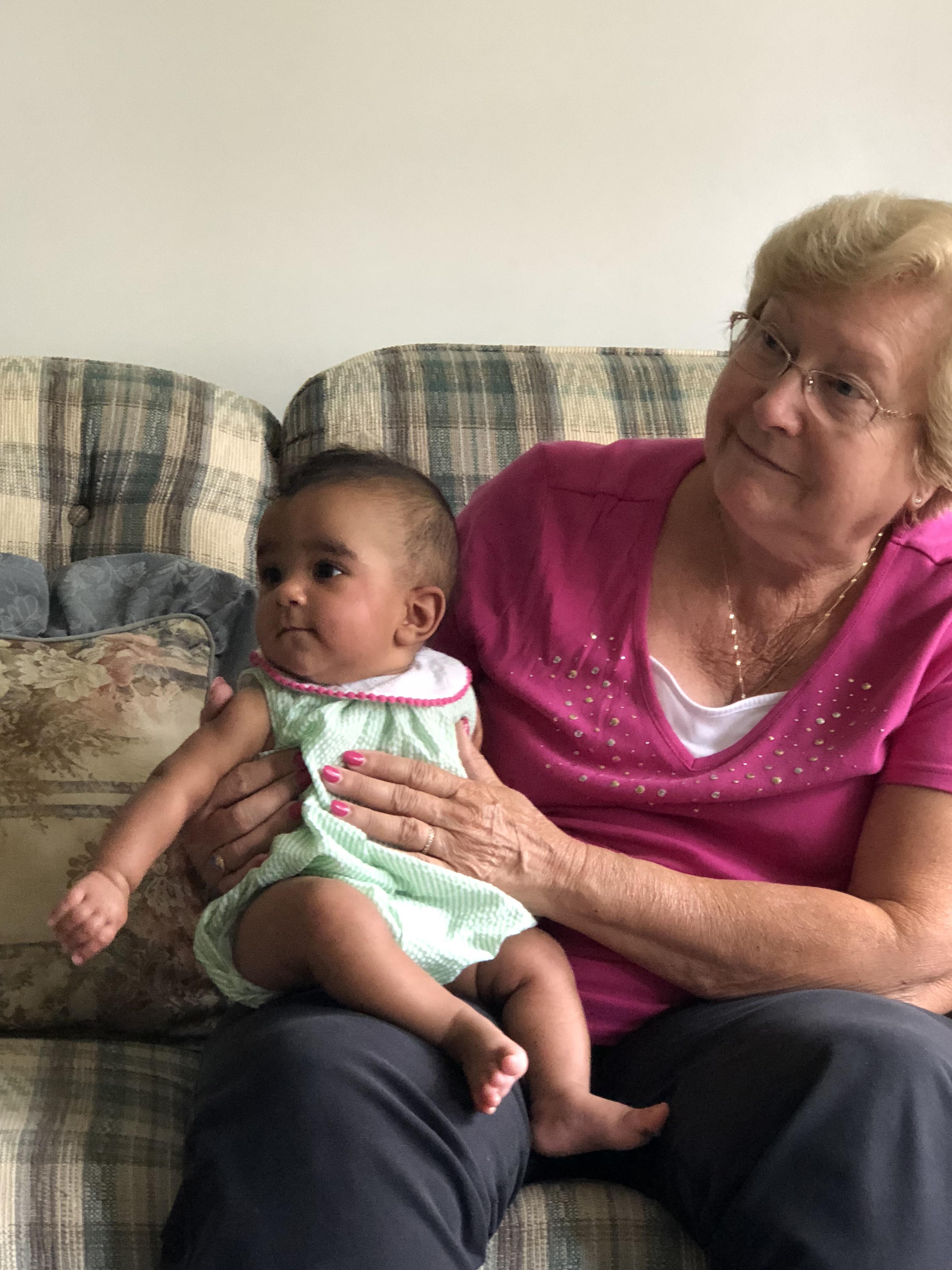 An elderly woman smiles while holding a baby on a couch in a comfortable living room.