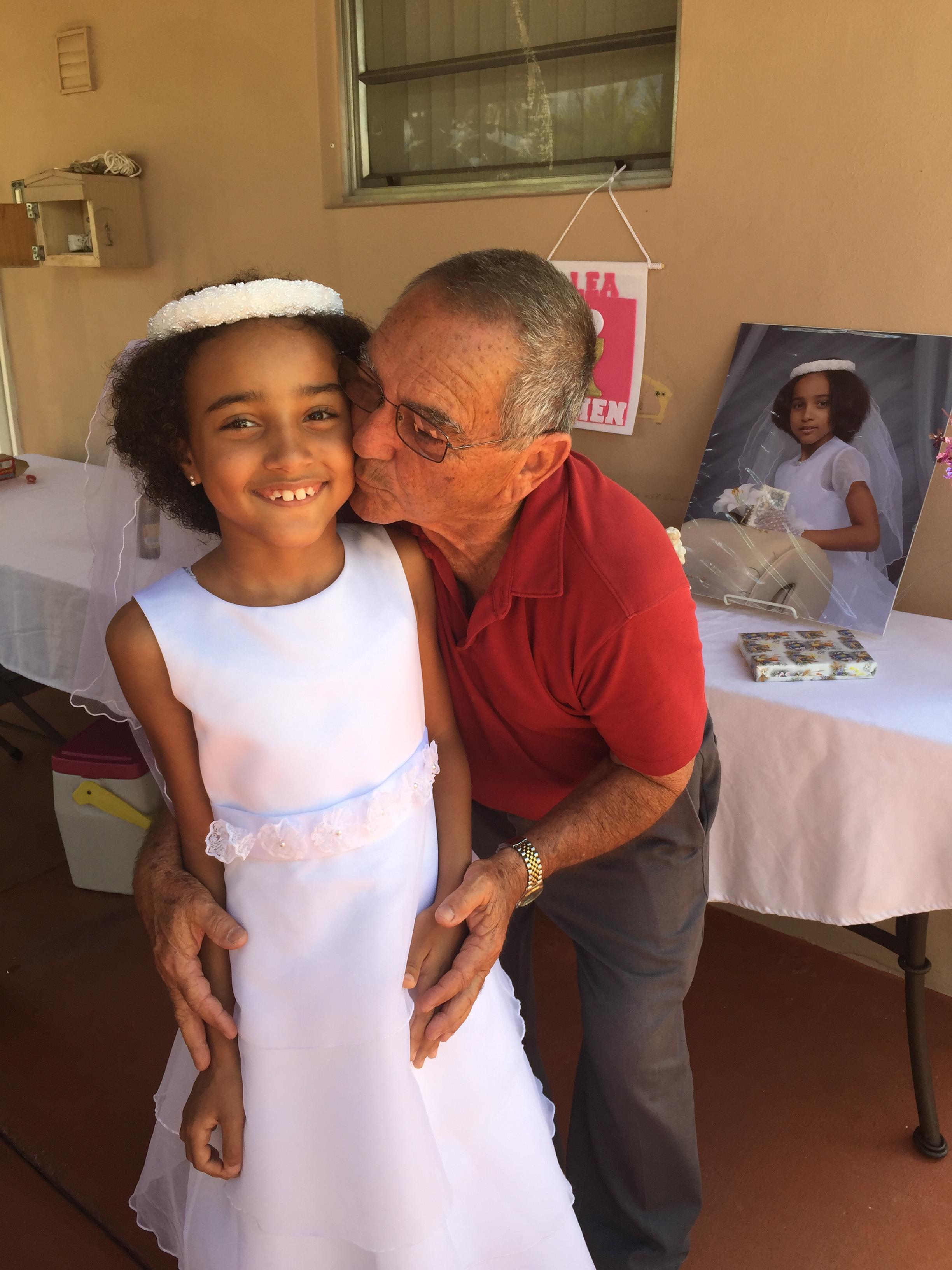 A grandfather kisses his smiling granddaughter in a white dress, celebrating together.