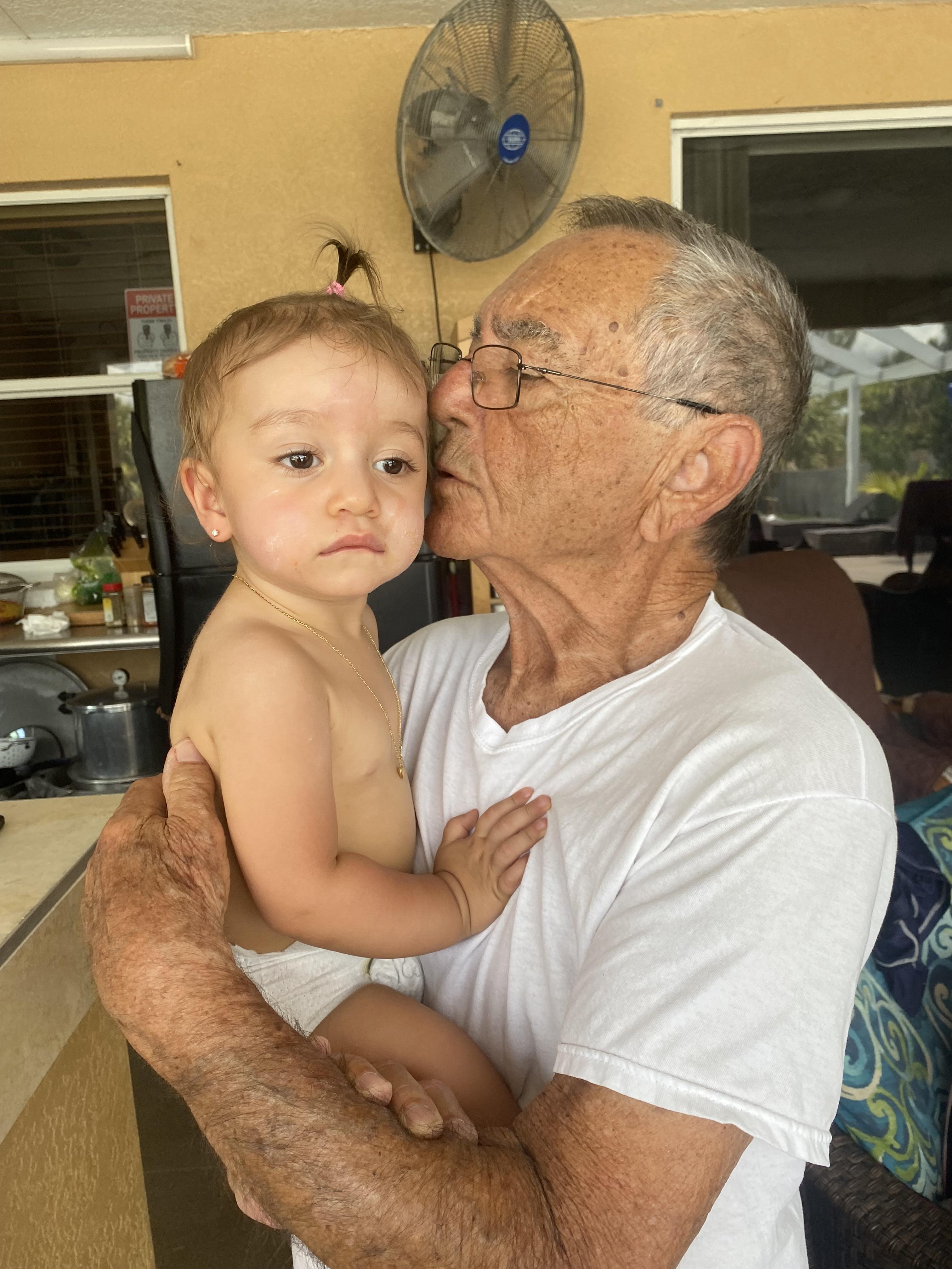 An elderly man gently kisses his grandchild indoors, sharing a heartwarming moment.