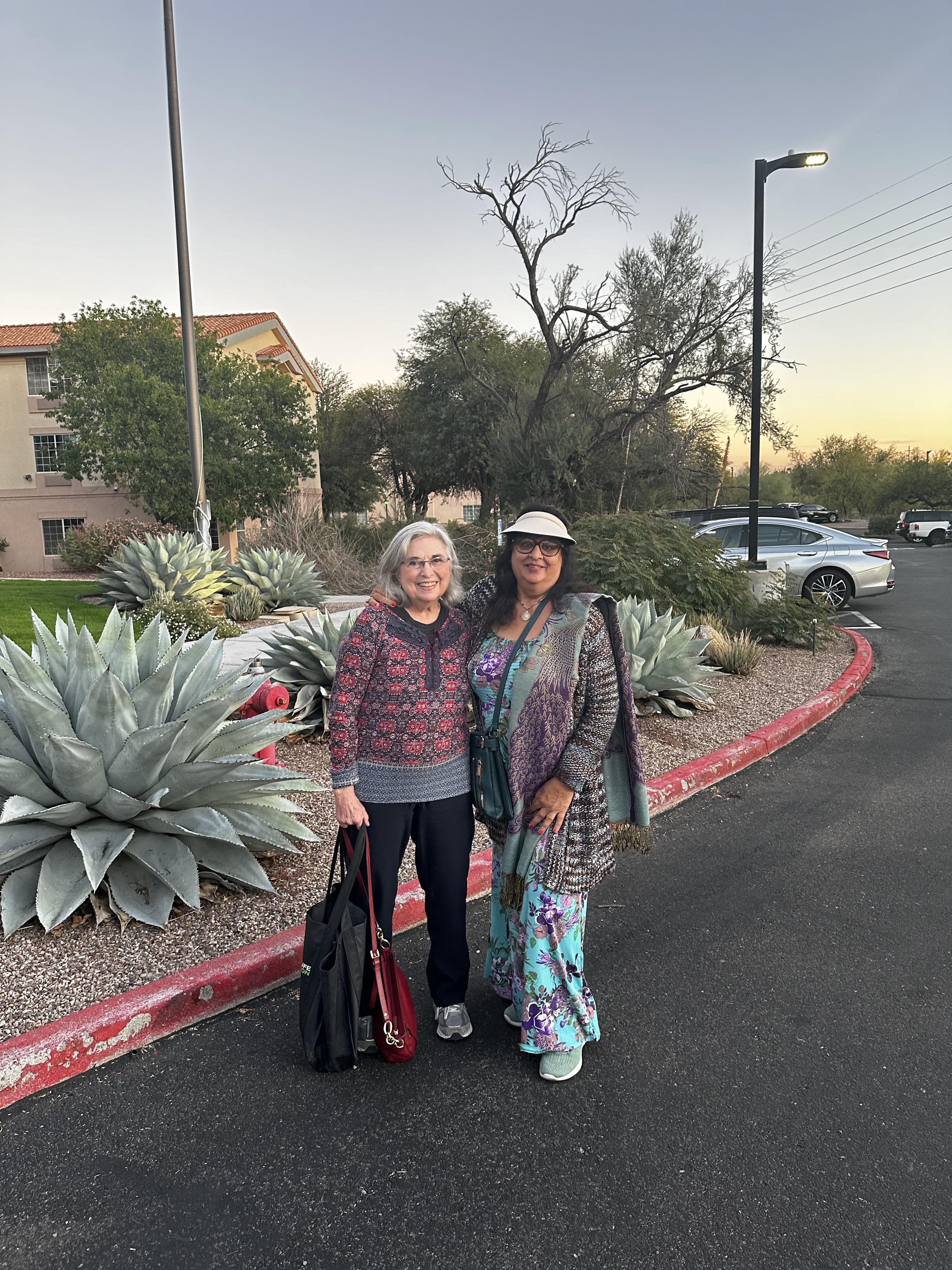 Two women stand together outside a hotel, smiling as dusk settles. Desert plants frame them.