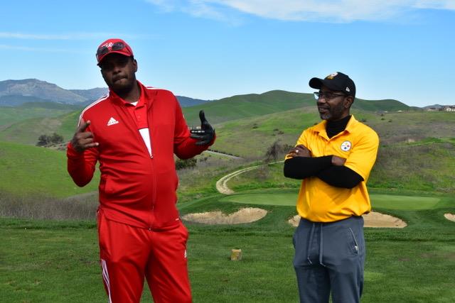 Two men engage in conversation while standing on a green golf course under a bright sky.