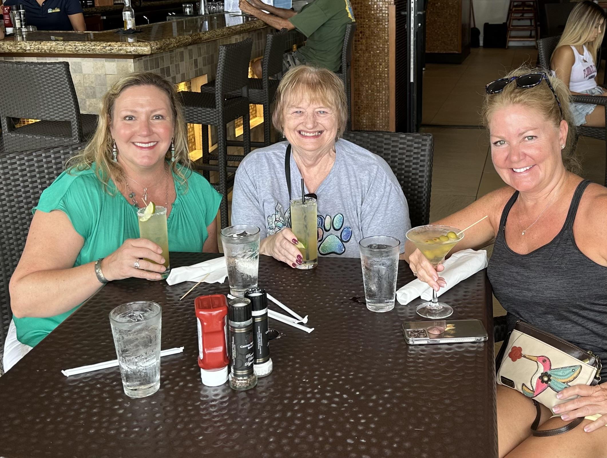 Three women gather at a restaurant table, smiling and sharing drinks in a vibrant atmosphere.