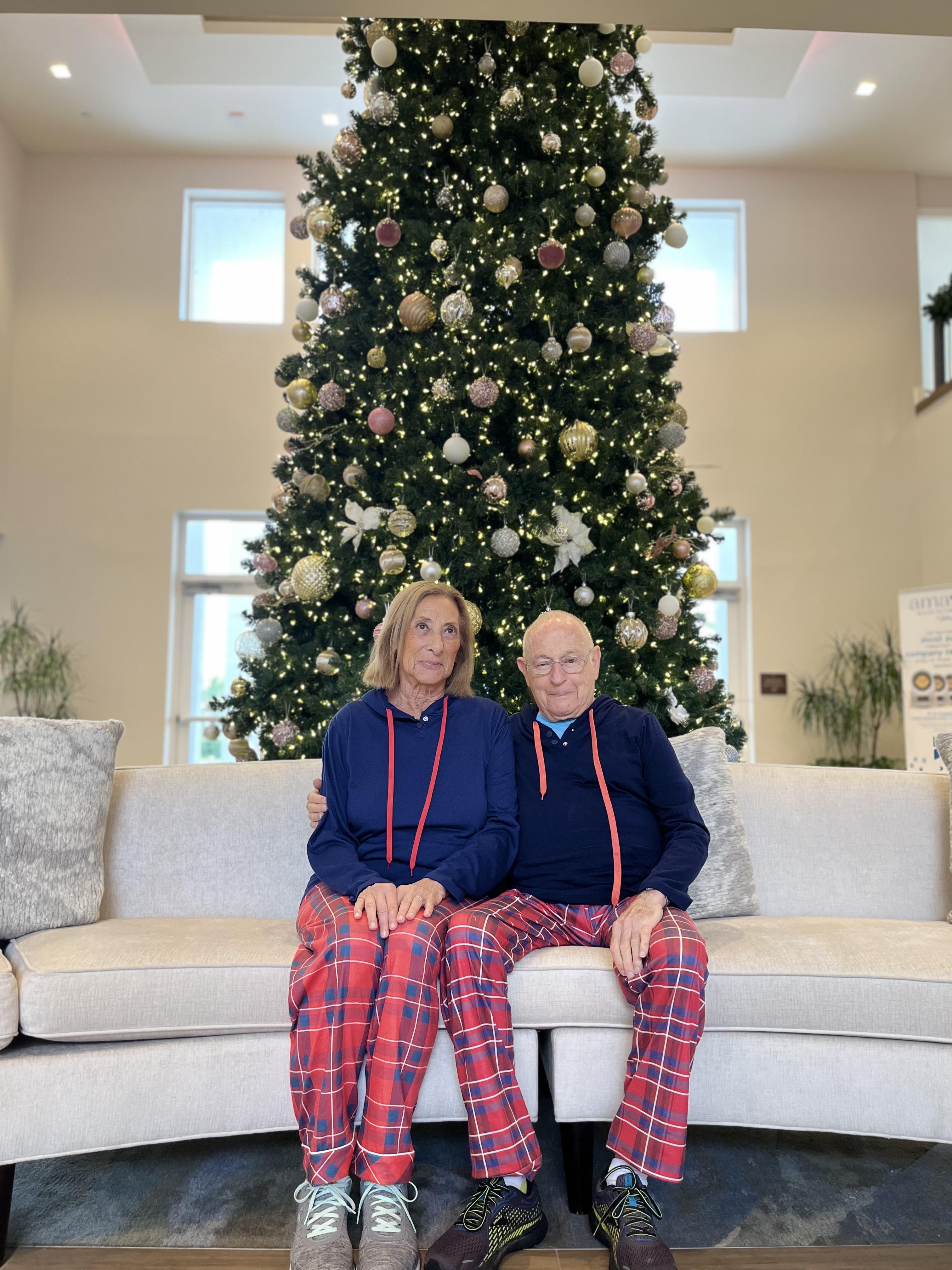 Couple in coordinating plaid pajamas relaxes on a sofa near a beautifully decorated Christmas tree.