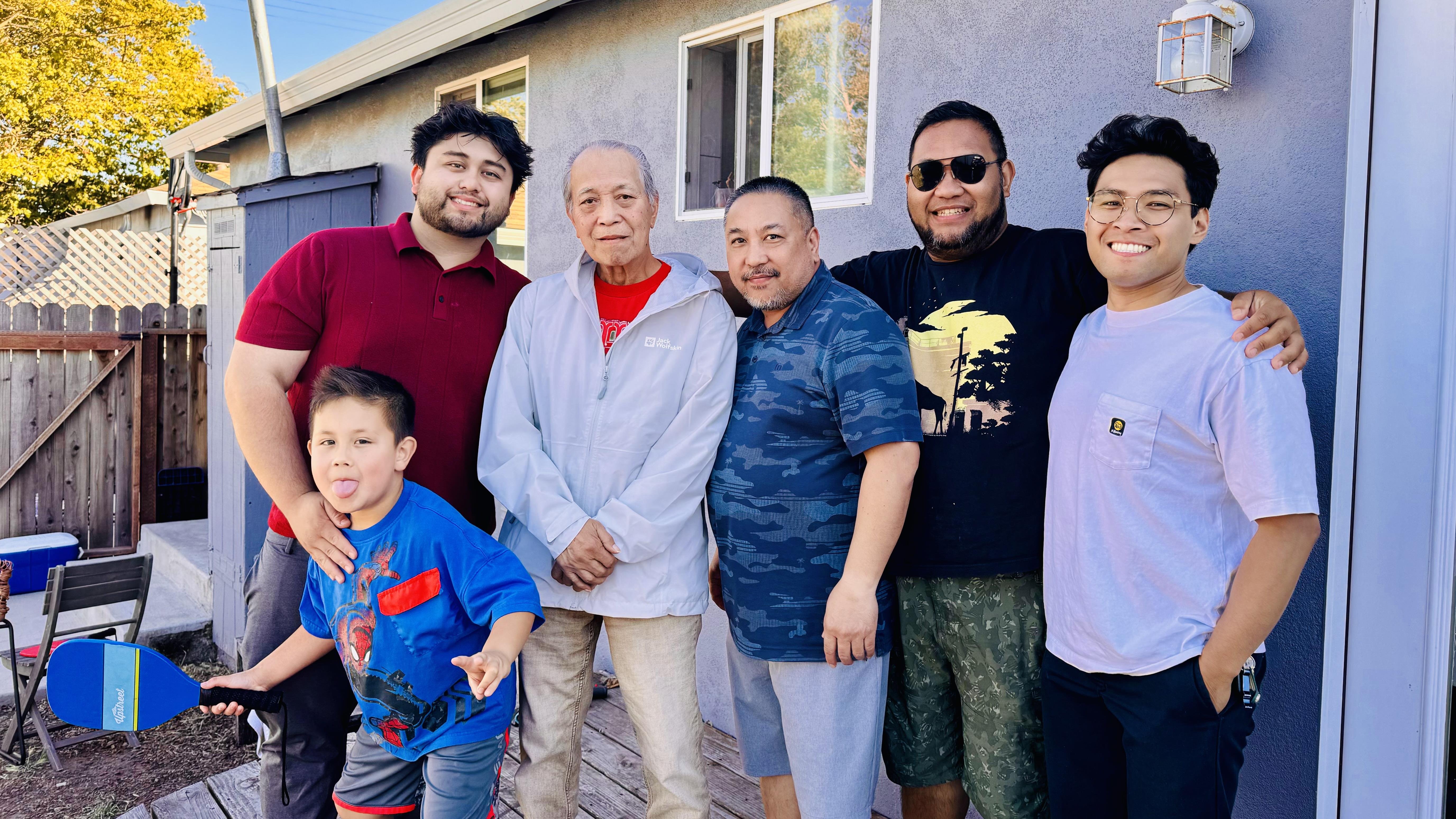 A group of five family members gather together smiling and enjoying a casual day outdoors.
