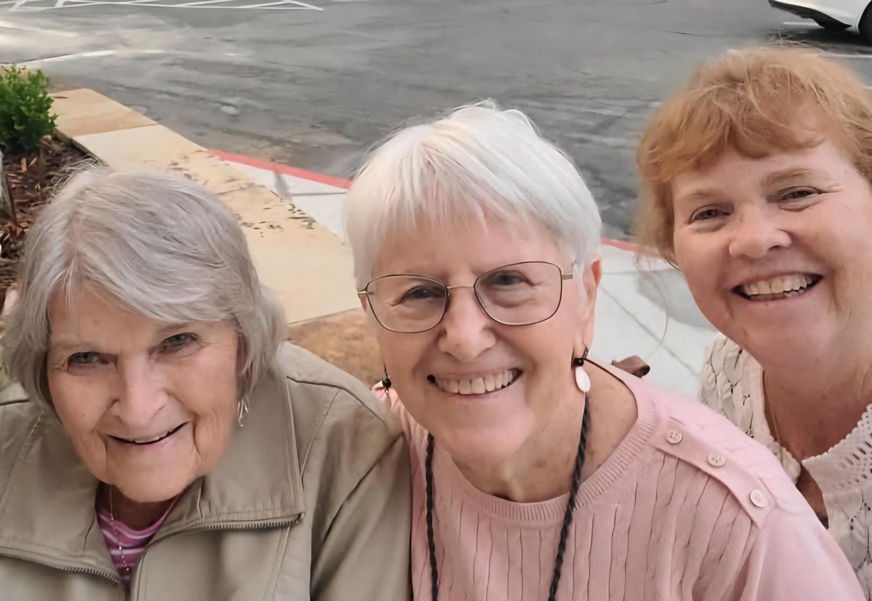 Three women smile joyfully while posing for a selfie in a sunny outdoor setting.