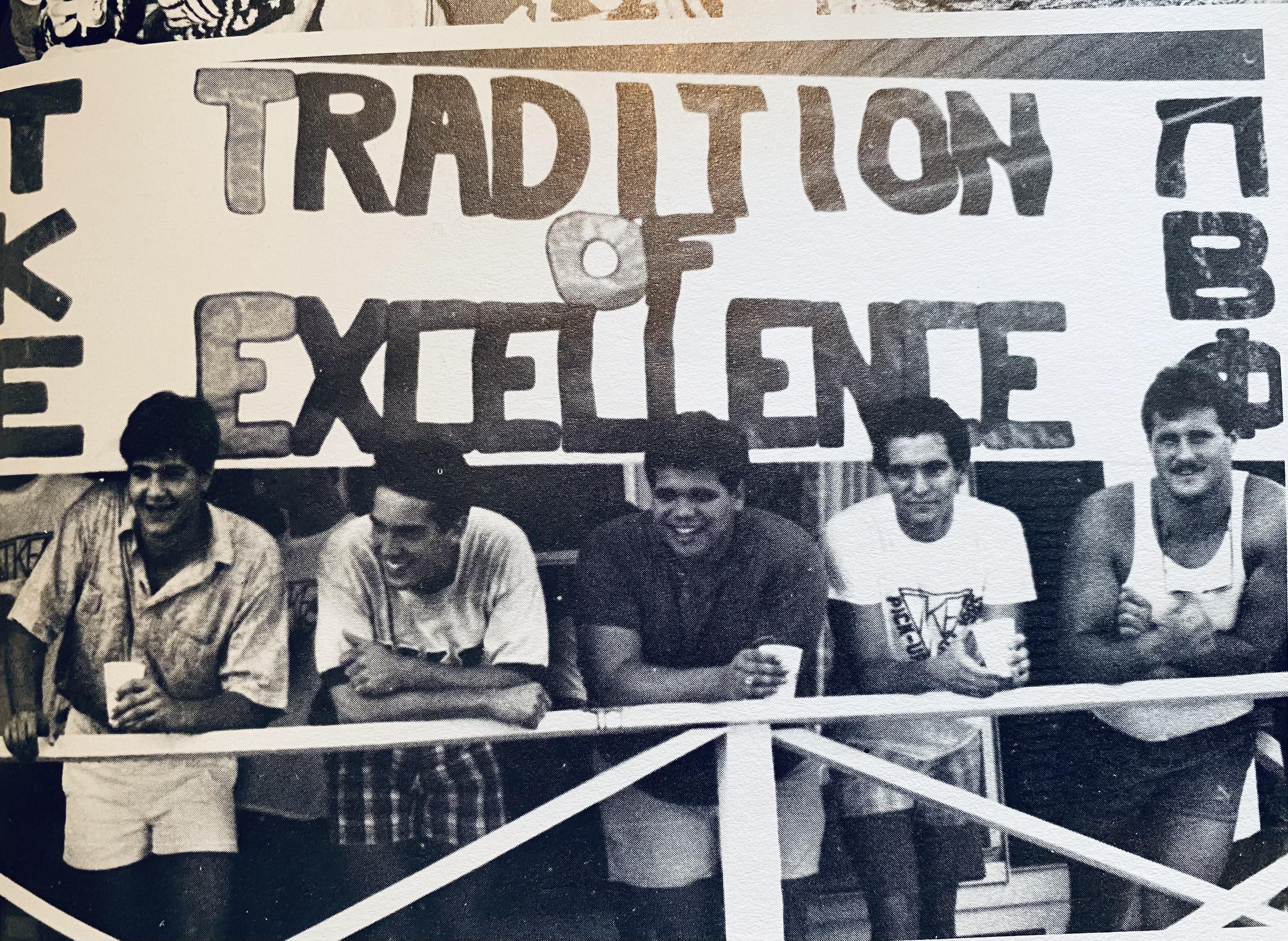 Friends showcase their camaraderie while holding drinks in front of a sign highlighting excellence.