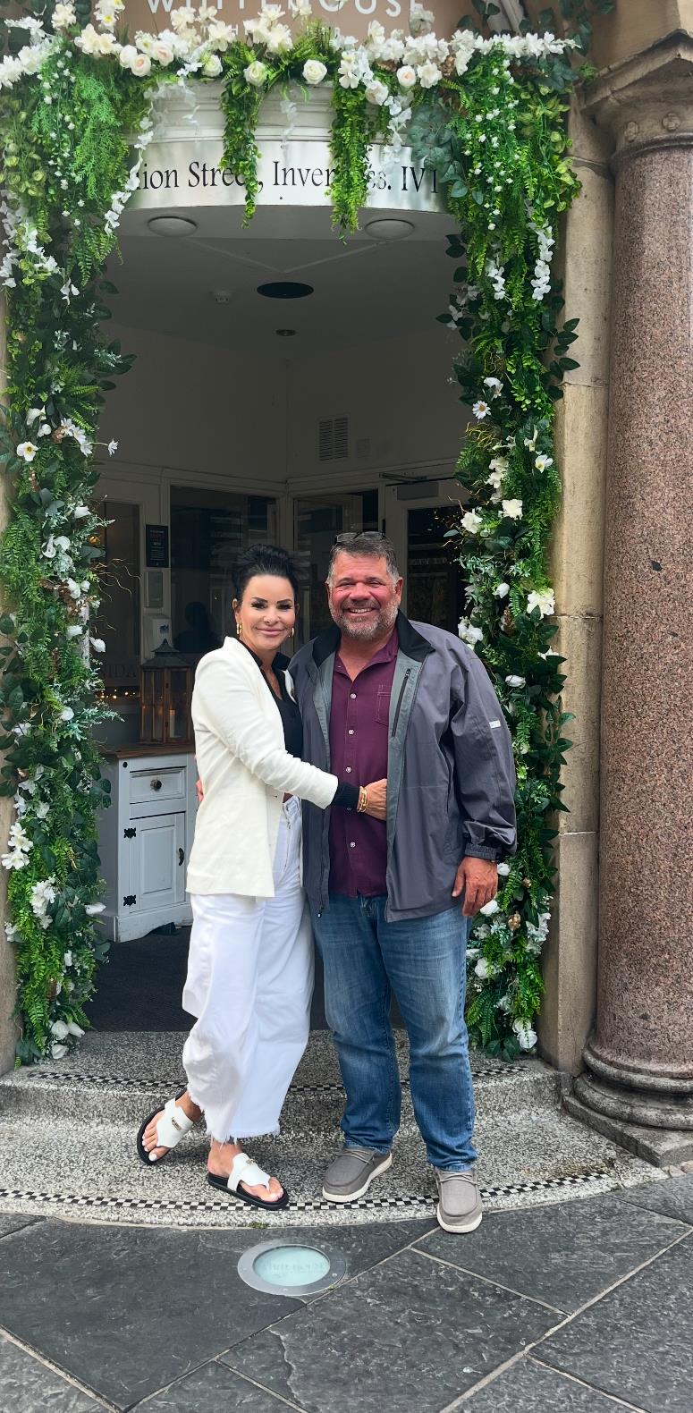 A couple stands together, smiling in front of a beautifully decorated entrance, enjoying a warm day.