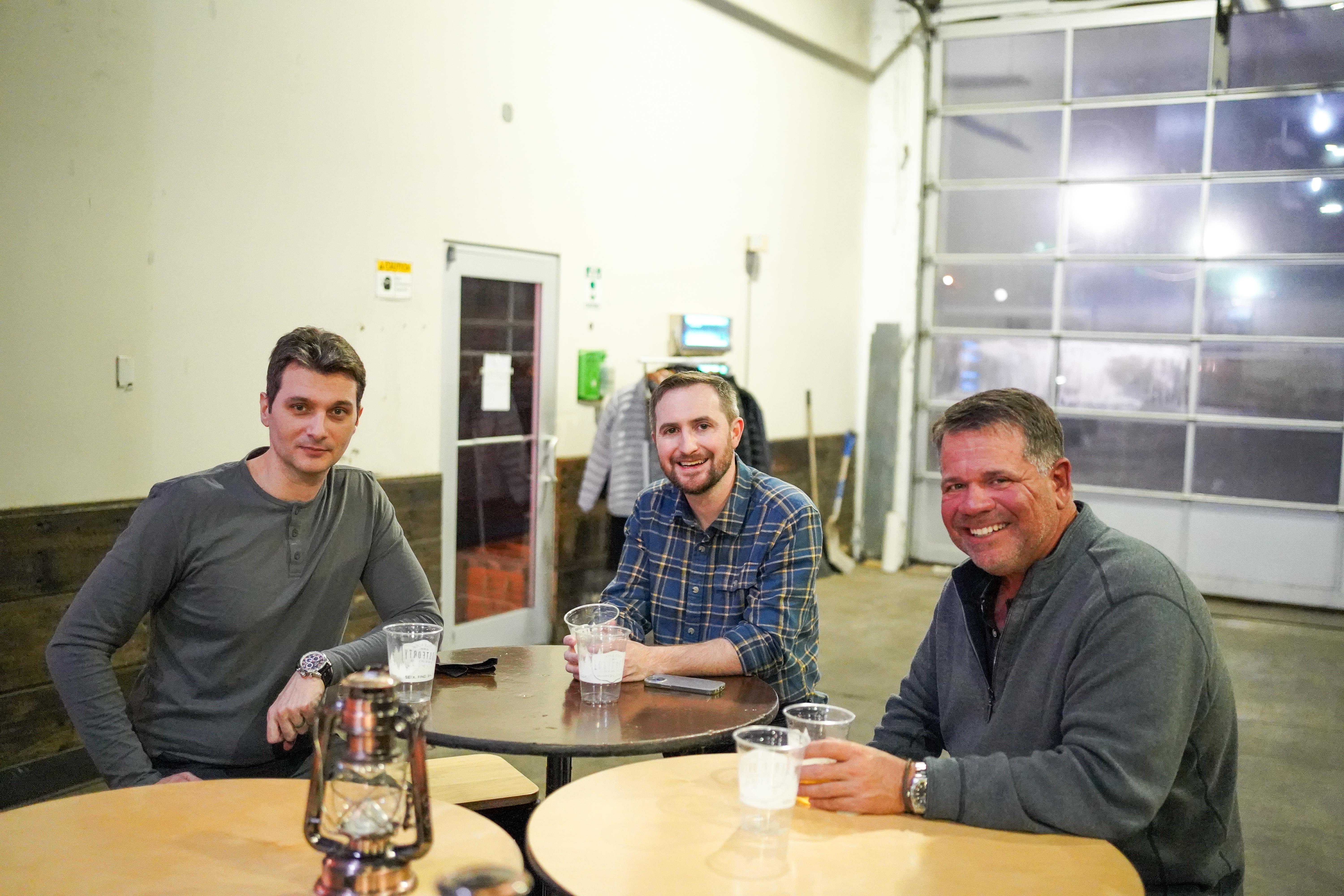 Three men engage in lively conversation while sipping drinks at a table in a spacious venue.