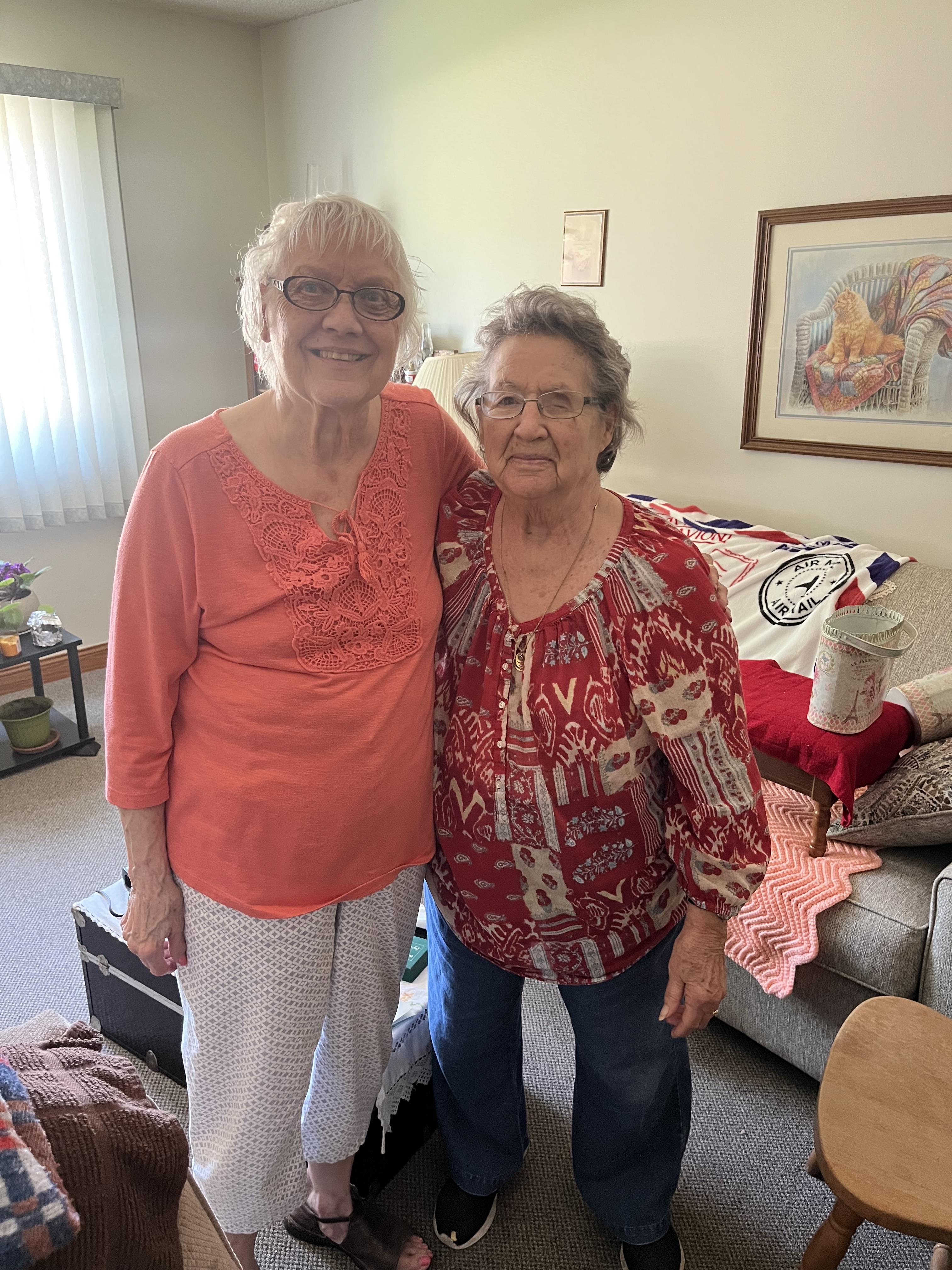 Two smiling elderly women stand together in a warm, inviting living room, celebrating friendship.