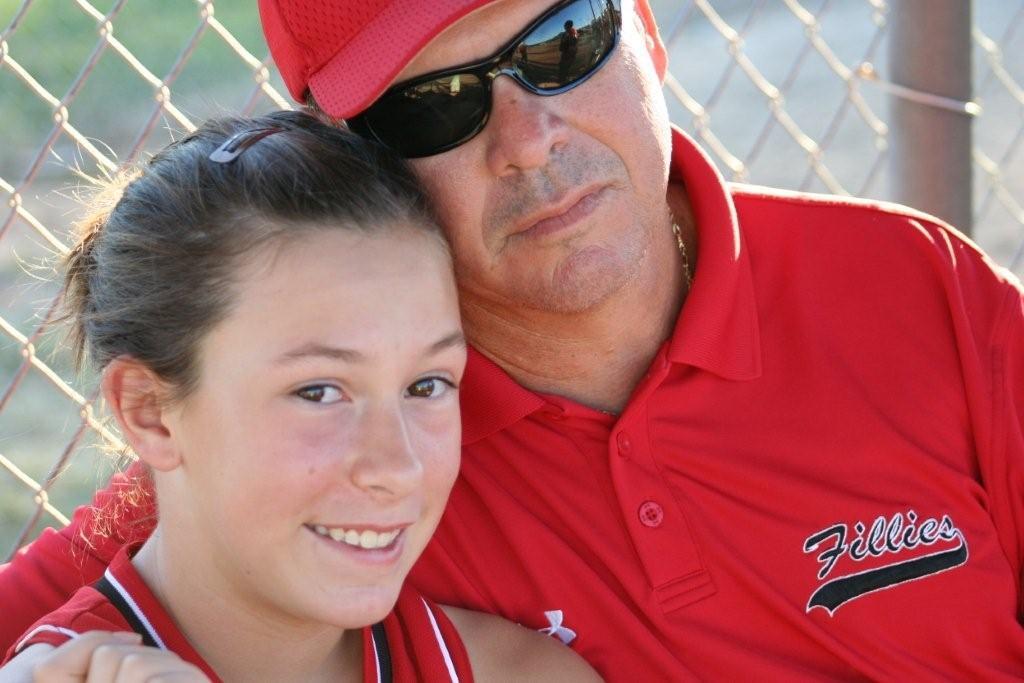 A young athlete smiles while leaning against her father at a sports event during summer.