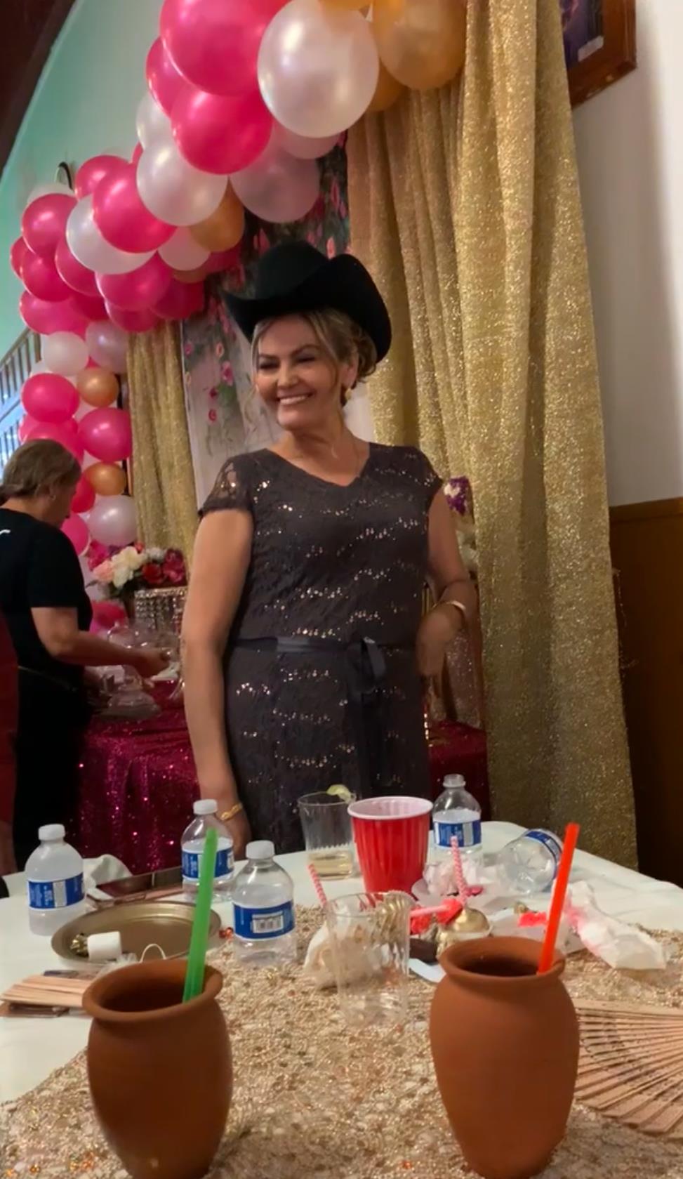 A cheerful woman stands confidently with a hat at a lively celebration adorned with balloons.