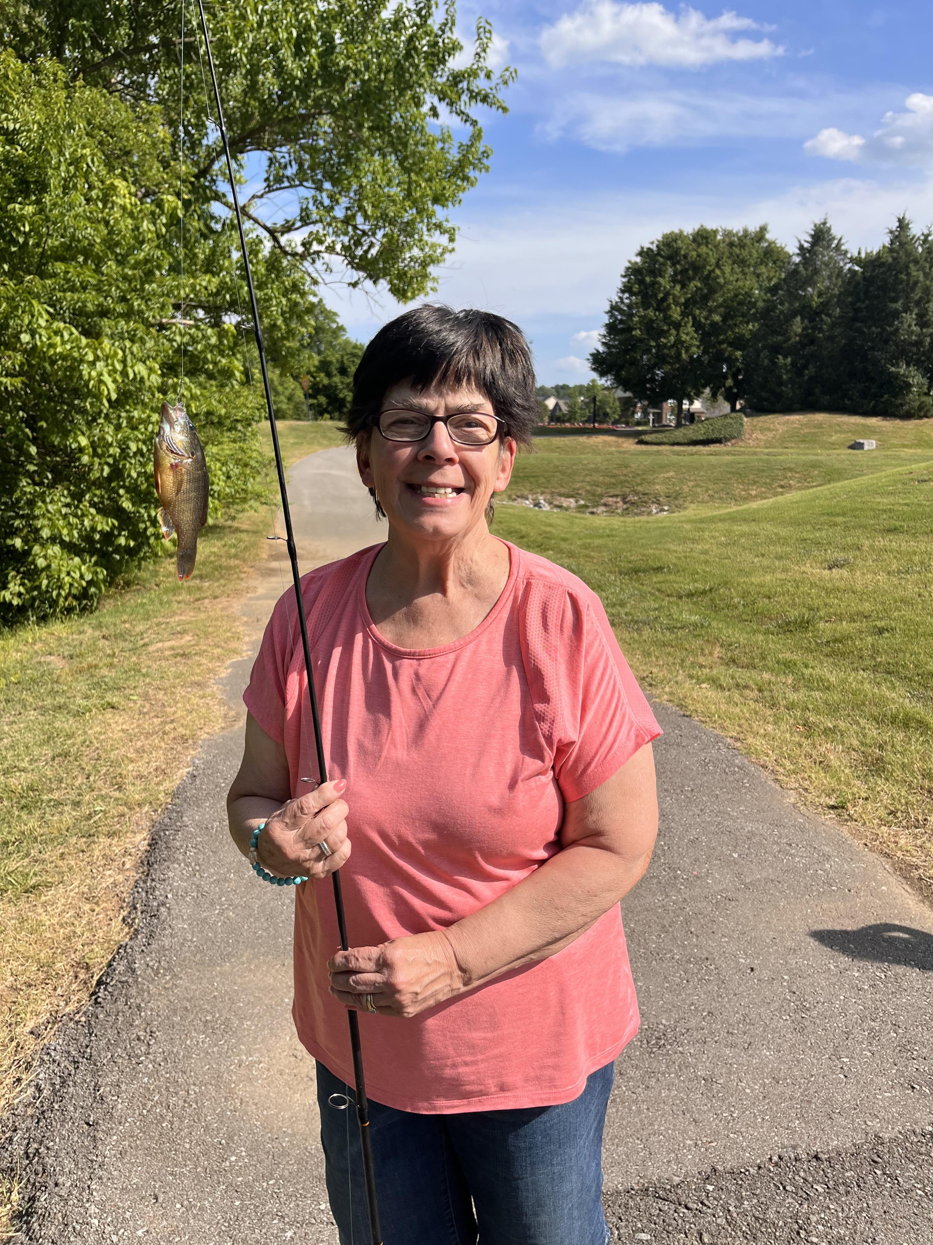 A woman smiles as she displays her freshly caught fish on a sunny day in a park.