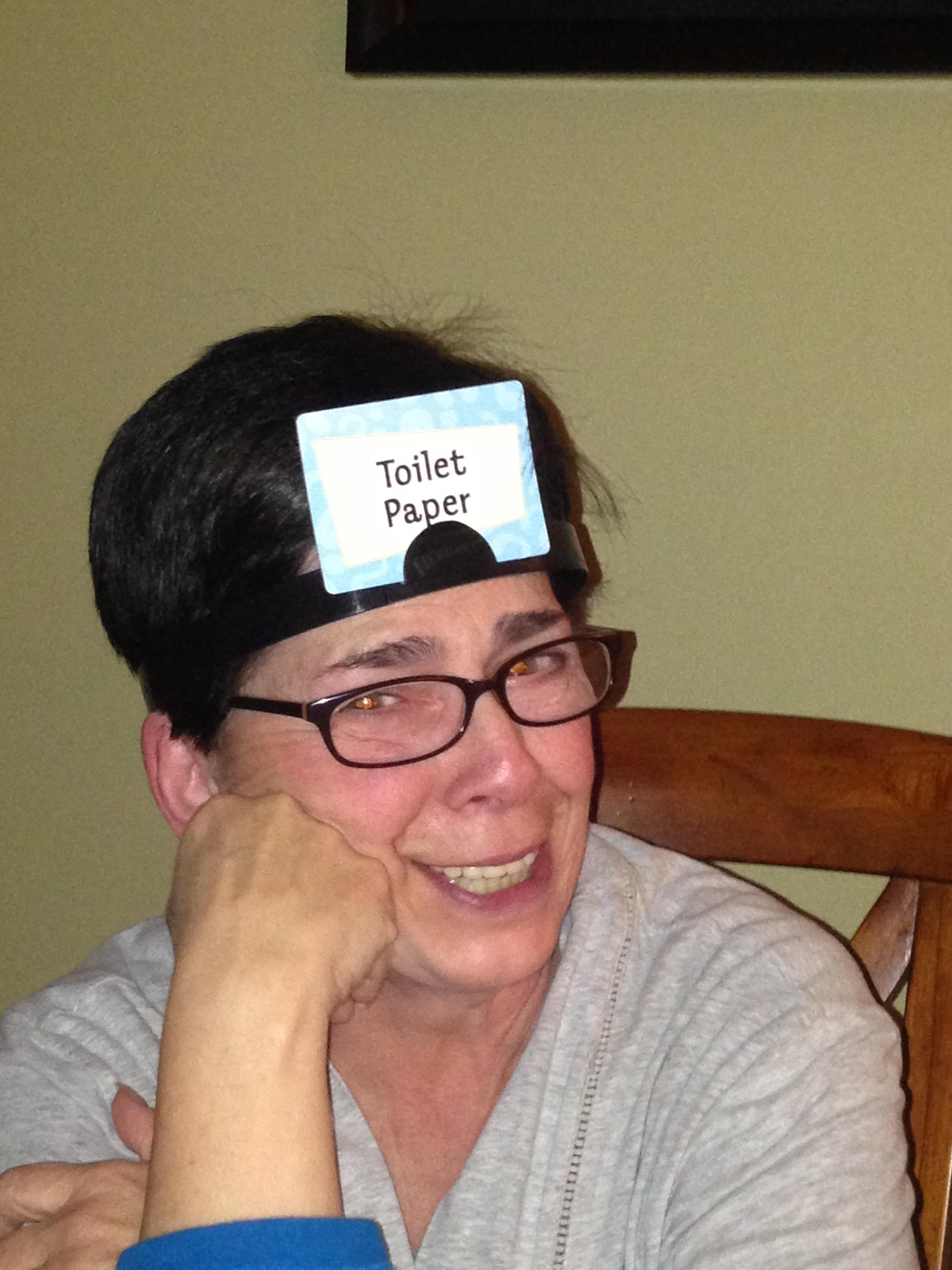 A woman enjoys a lighthearted game at home, wearing a card that says toilet paper on her forehead.