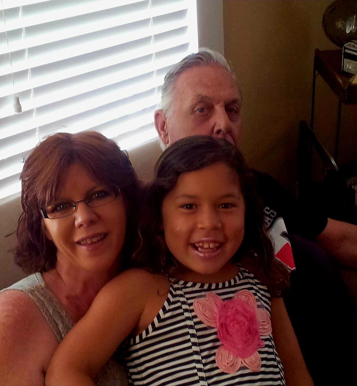 A young girl beams with happiness while sitting with two adults in a comfortable indoor environment.