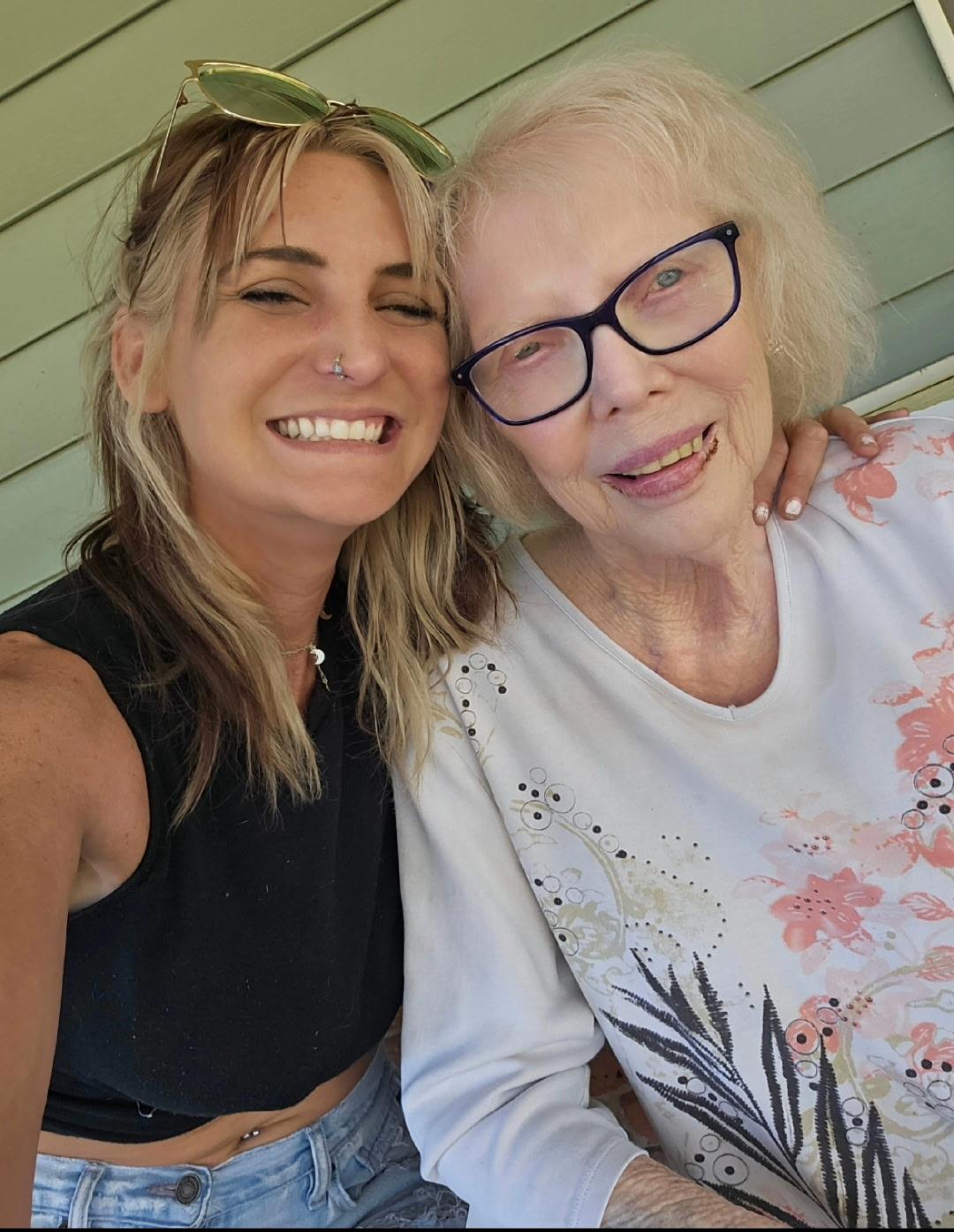A young woman and an elderly lady smile together on a porch, enjoying a sunny summer day.