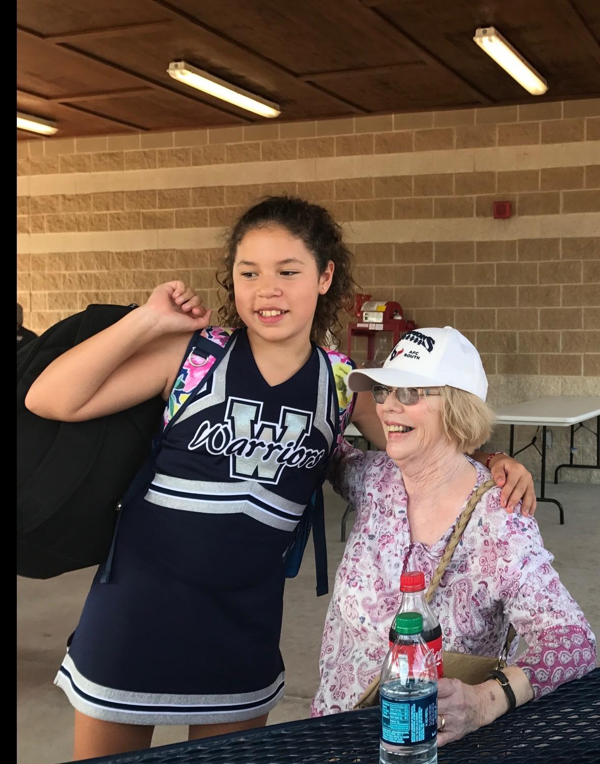A cheerful young cheerleader stands beside her grandmother at a sports event, both smiling happily.