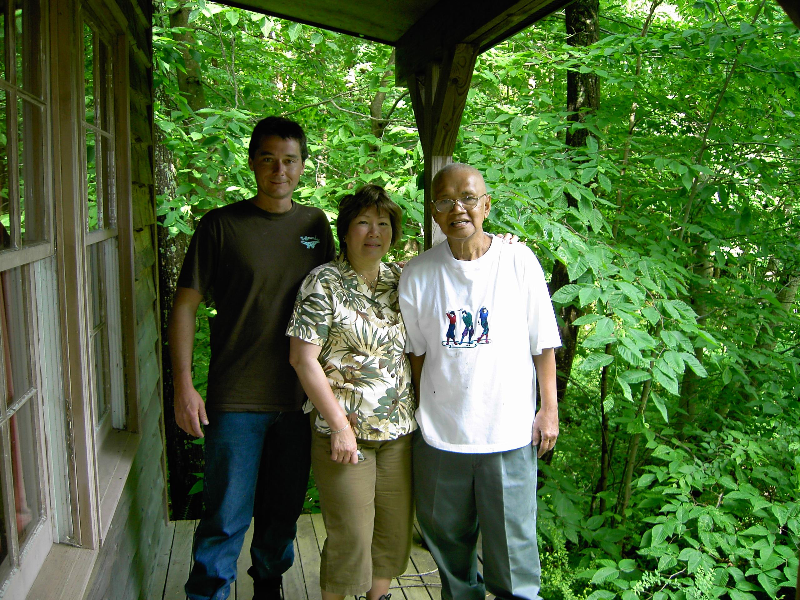 Three family members enjoy time together on a wooden porch surrounded by green foliage.