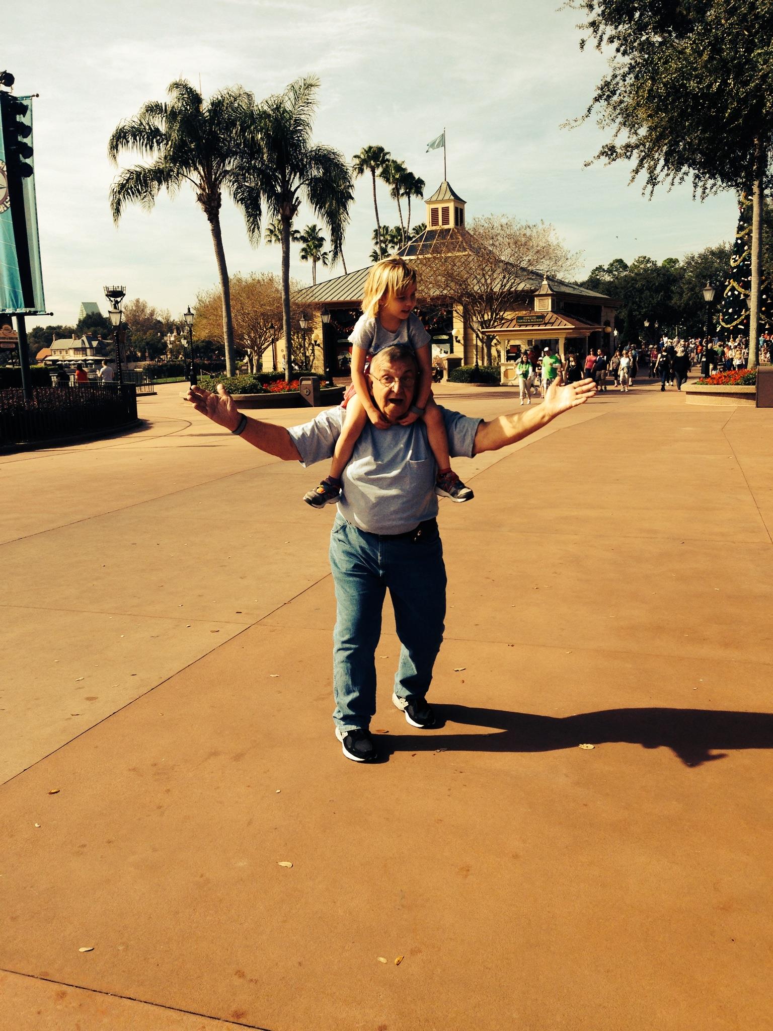 A man carries a child on his back while walking through a lively amusement park under clear skies.