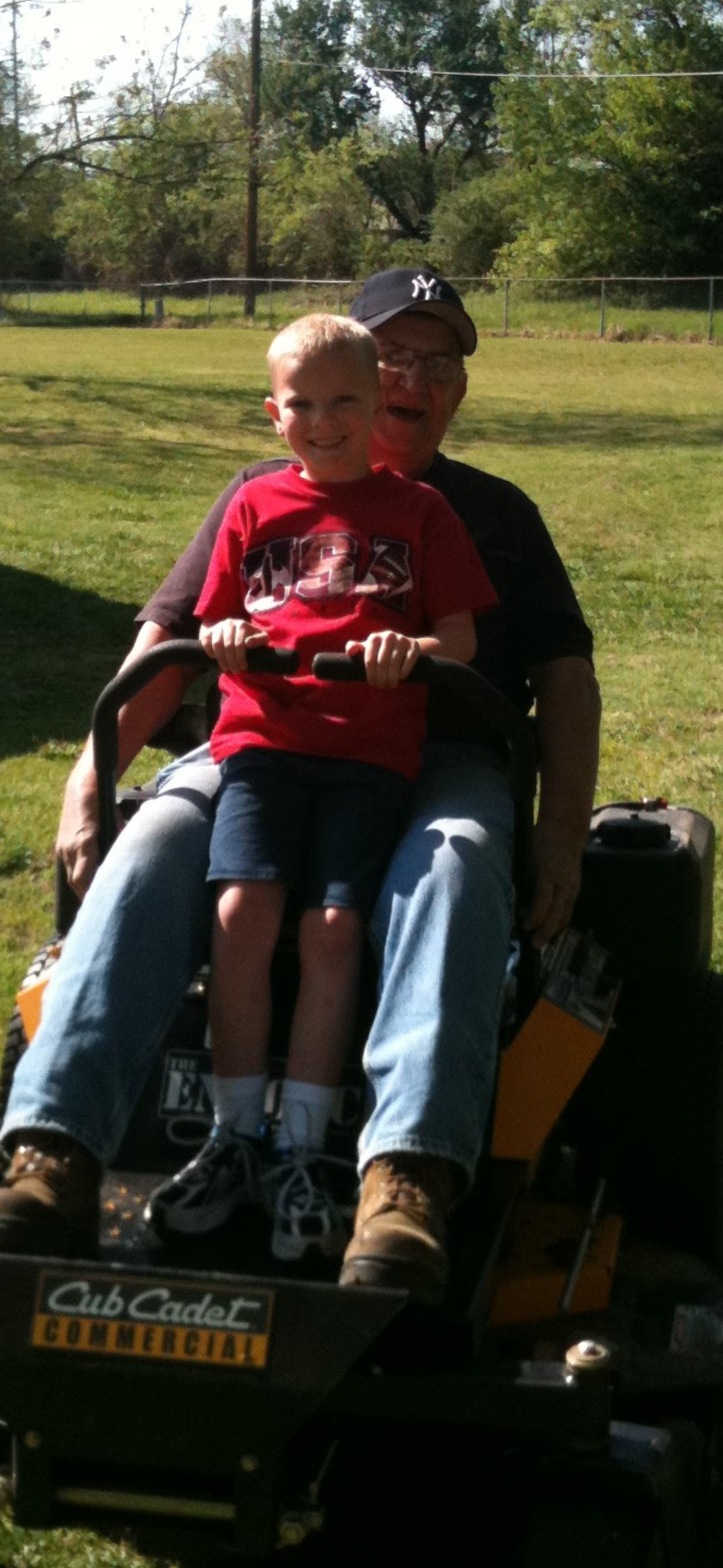 A man and a young boy share smiles while sitting on a riding lawnmower, enjoying their time outside.