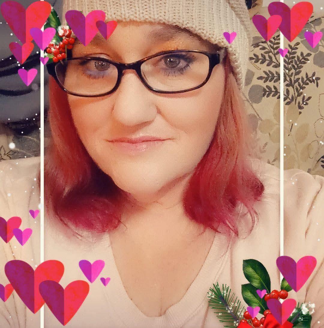 A woman with pink-tinted hair enjoys a moment indoors, smiling while wearing a knit hat and glasses.