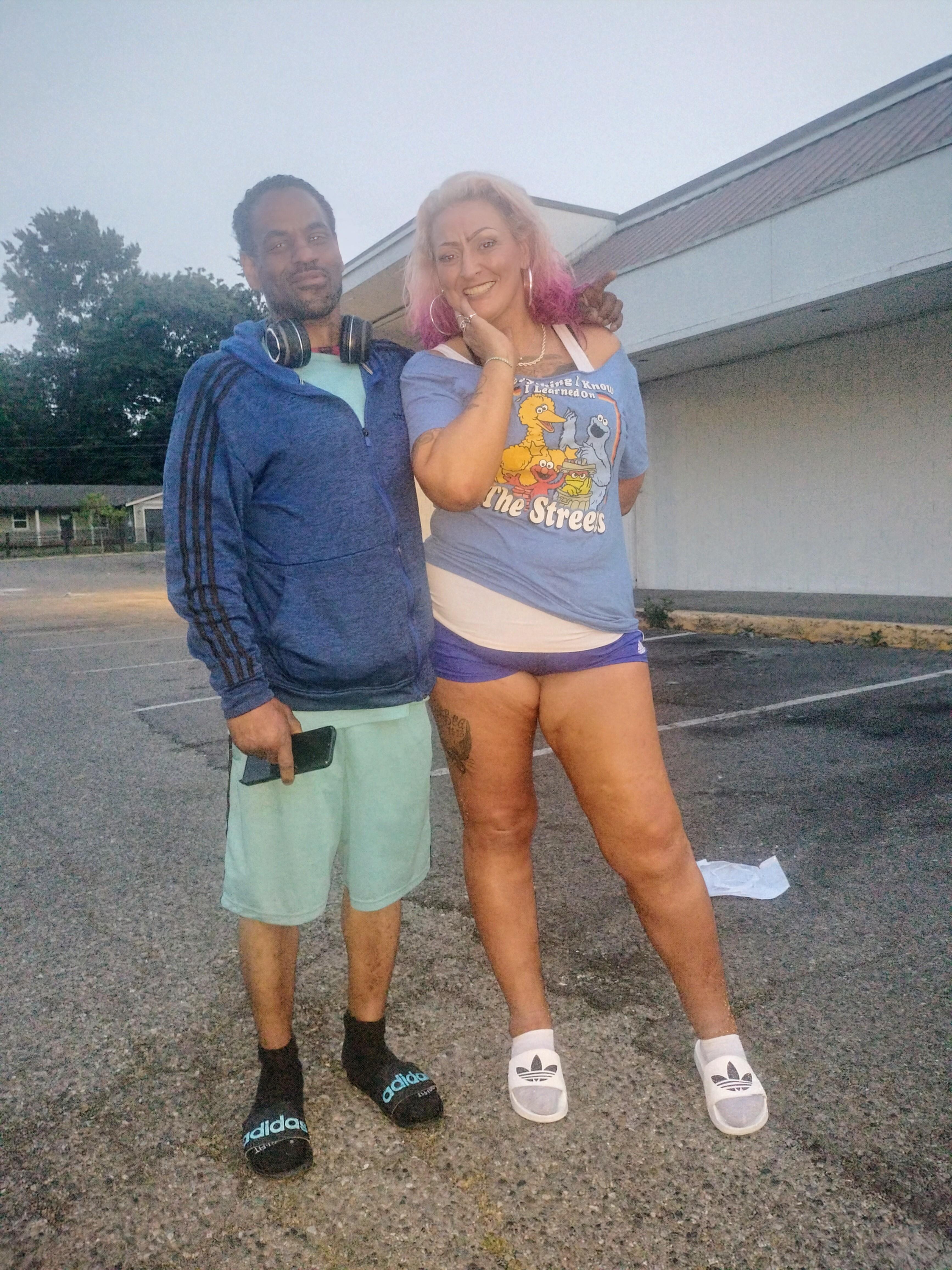 A couple stands together outside a shopping area at dusk, dressed casually.