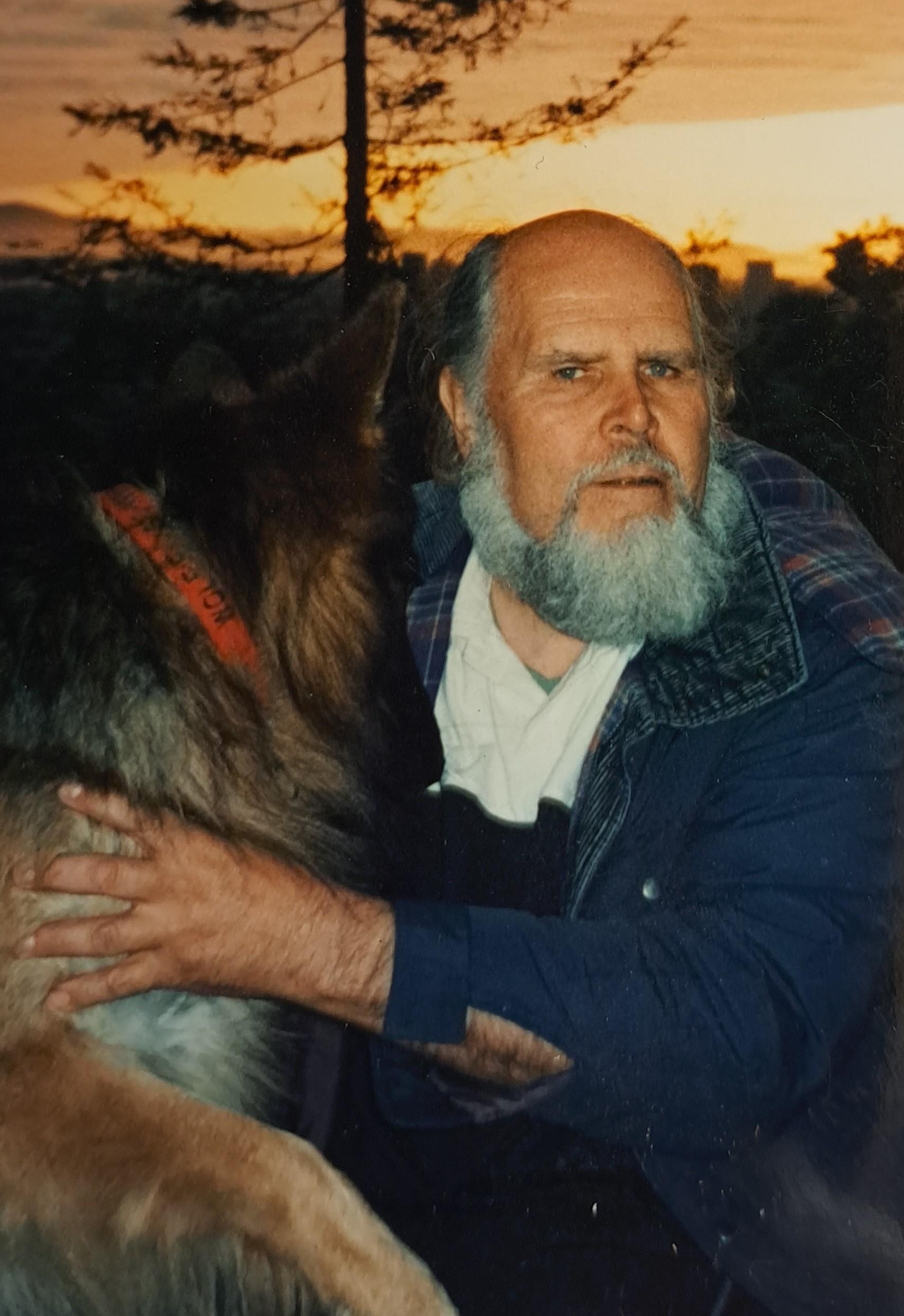 An elderly man with a beard lovingly holds his large dog close while enjoying a beautiful sunset.