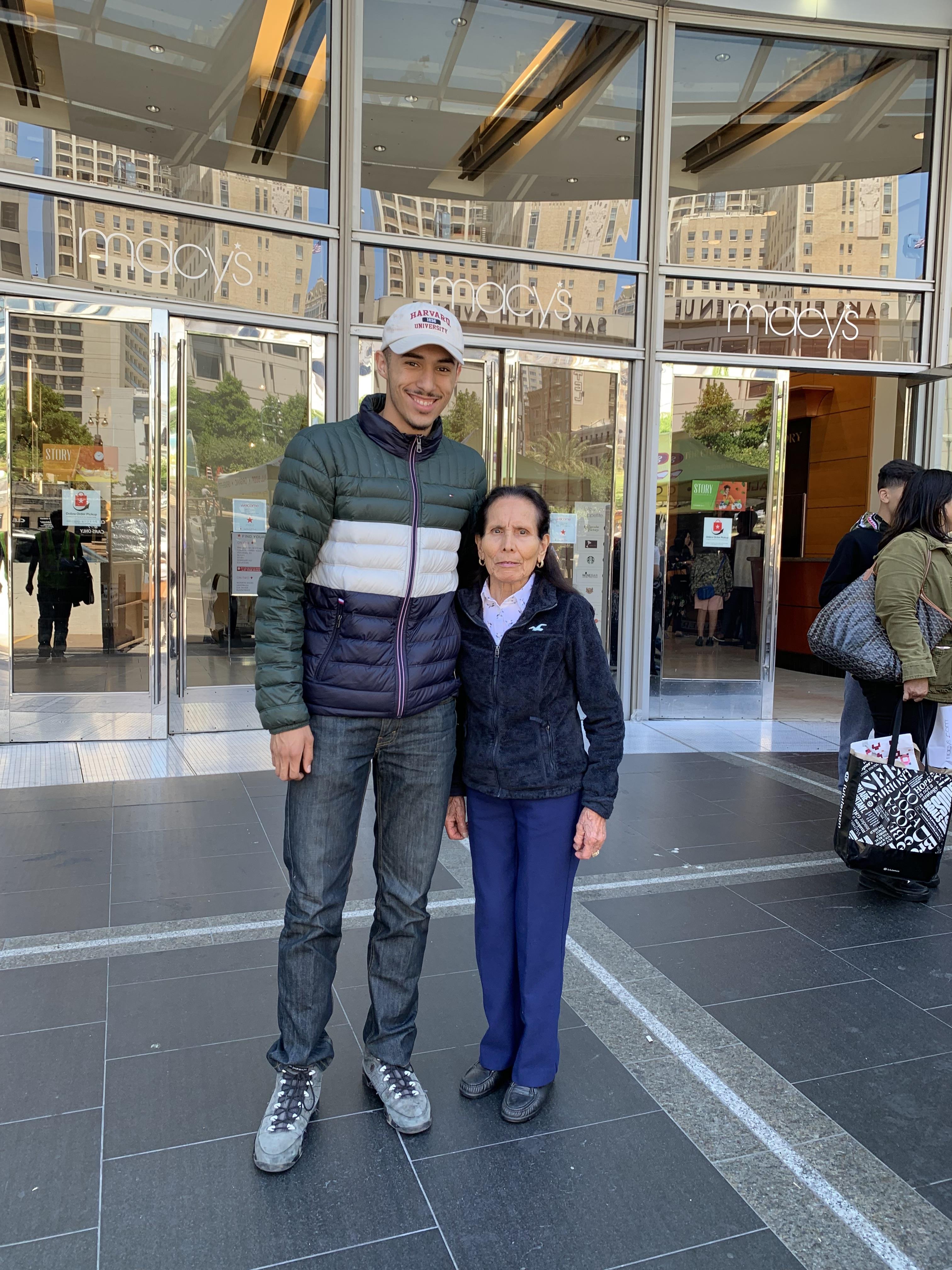 A young man and an older woman smile as they stand outside a bustling department store entrance.