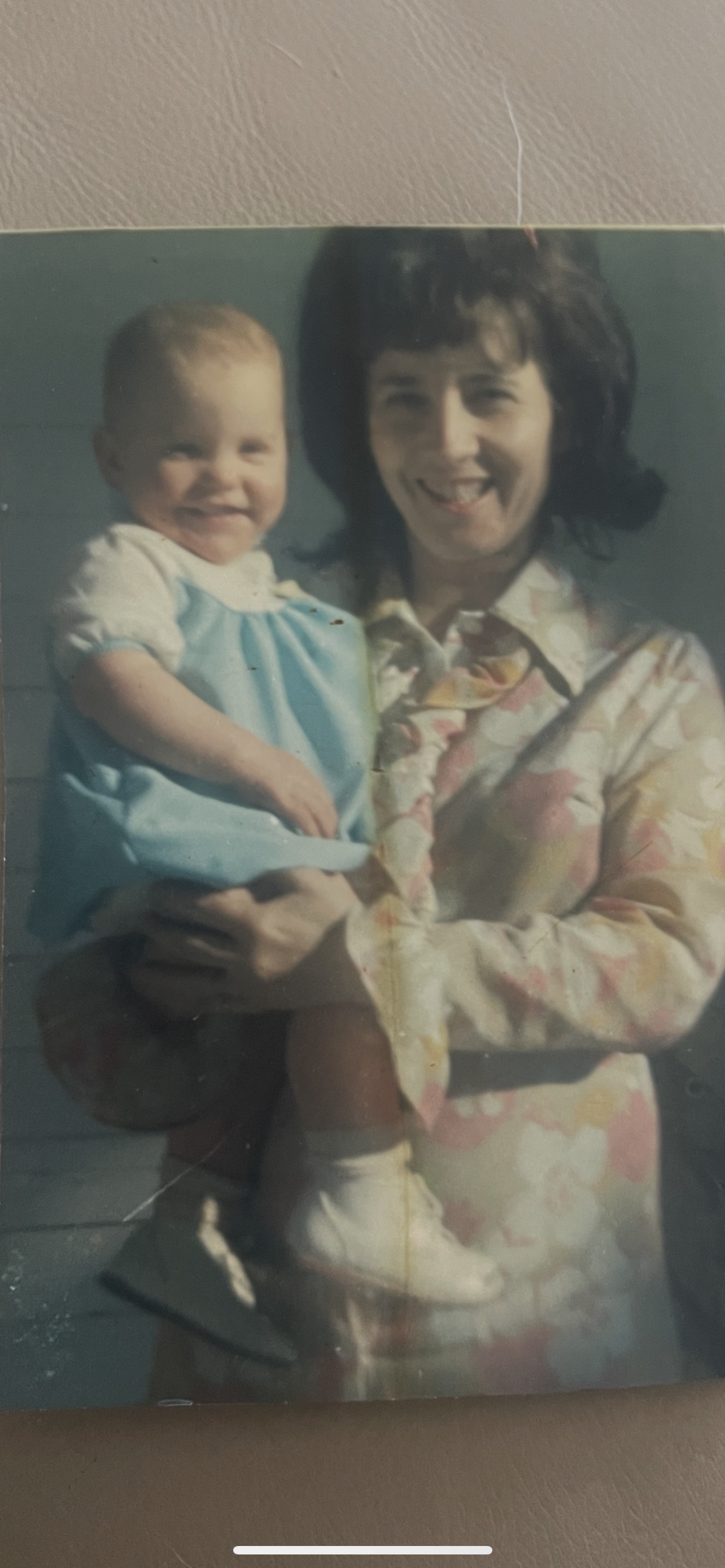 A cheerful woman wearing a floral dress holds a smiling baby dressed in blue.