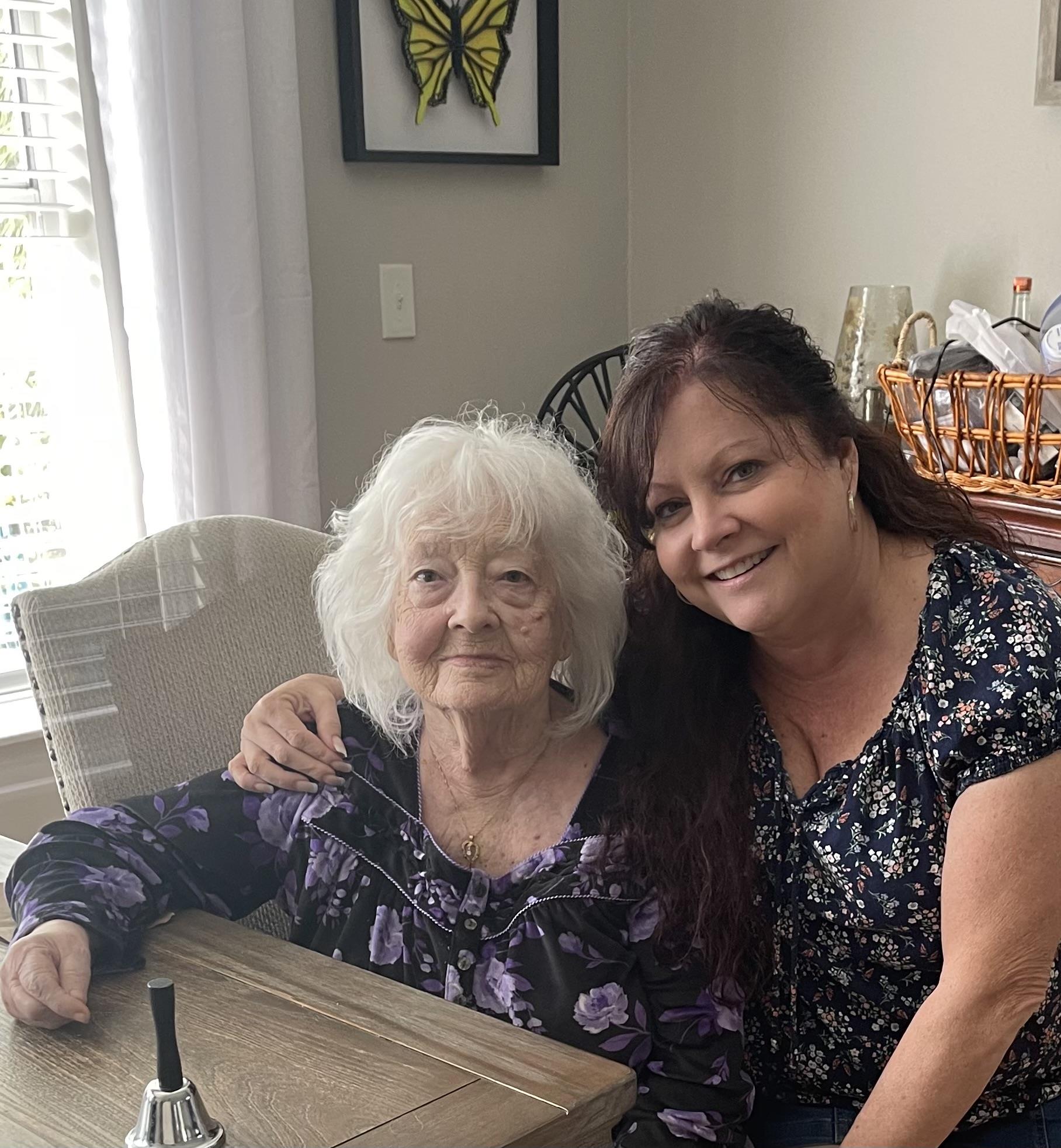Elderly woman with white hair and caregiver pose together in a bright, inviting living room setting.