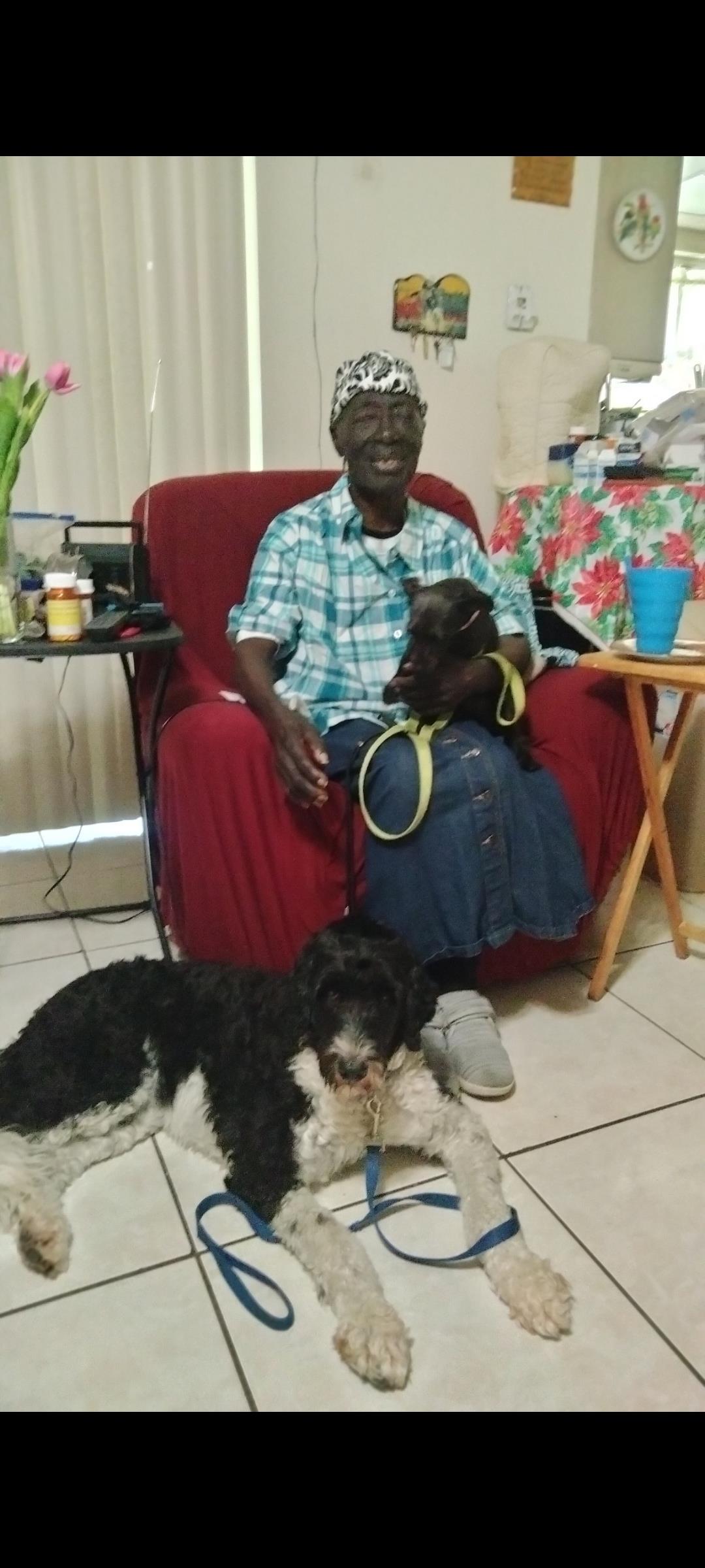 An elderly woman smiles while holding a dog in her lap, with another dog resting on the floor.
