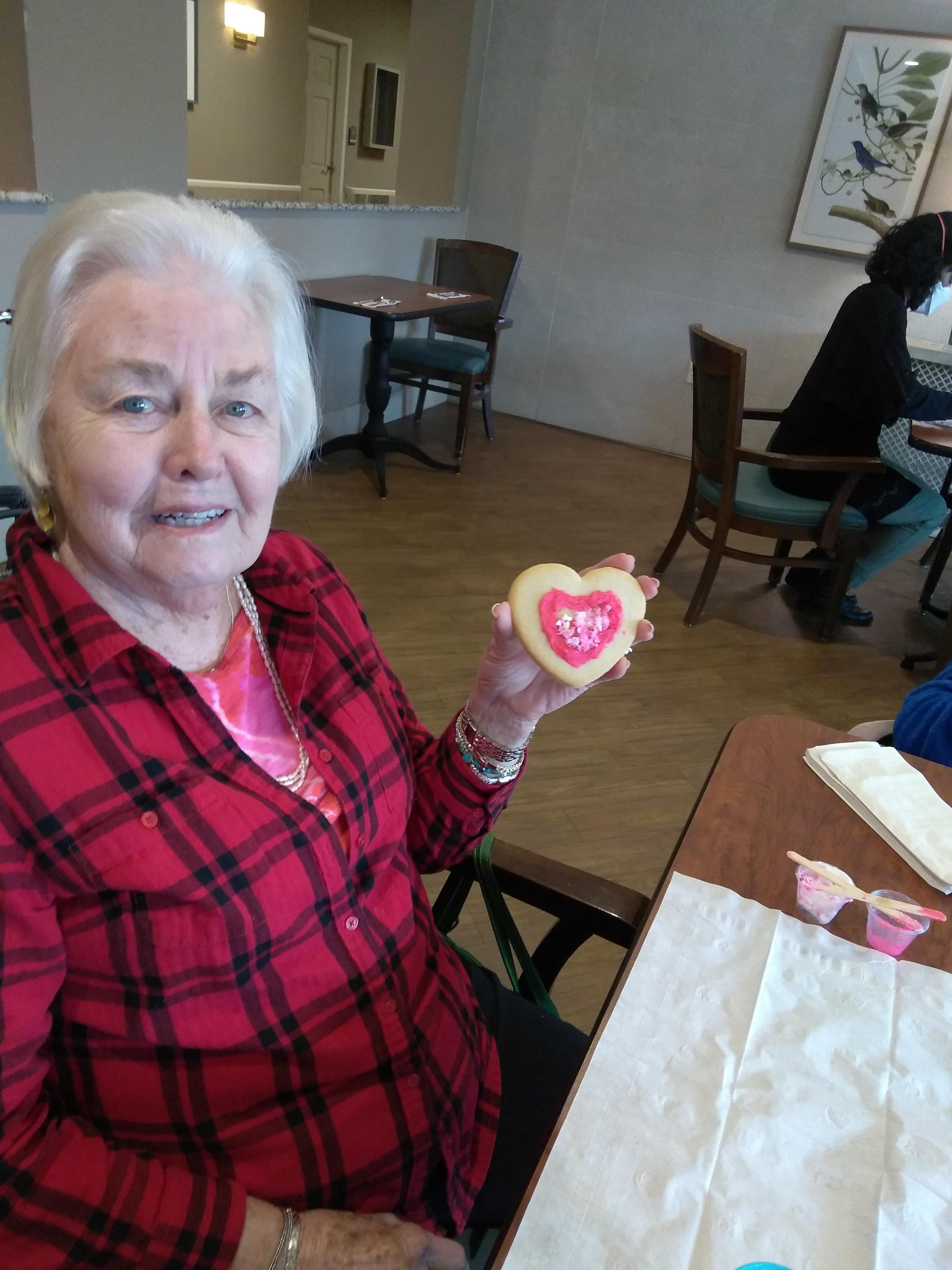 An elderly woman in a plaid shirt shows a heart-shaped cookie she decorated.