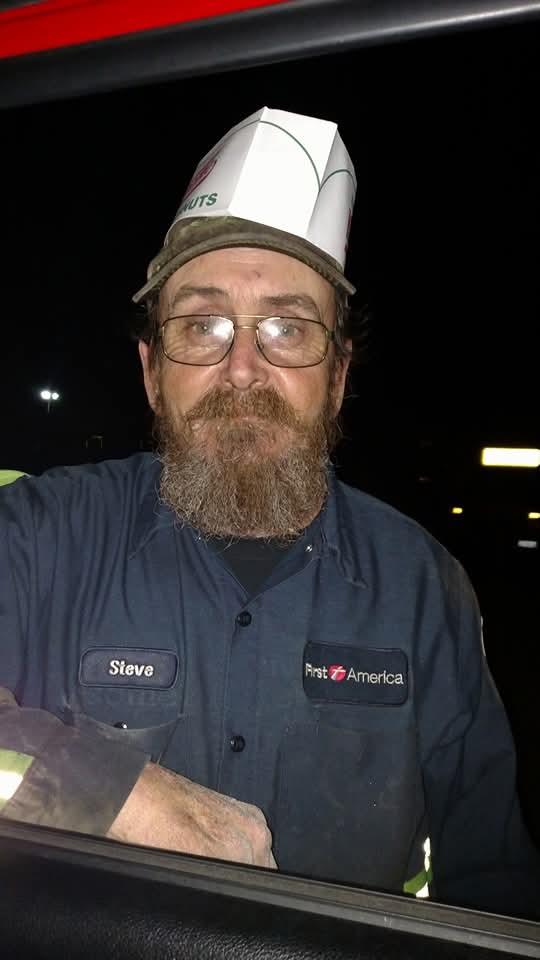 A man in a blue work uniform and glasses smiles from the window of a parked car during nighttime.