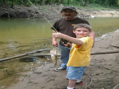 A young boy proudly holds a caught fish while fishing beside an adult on a riverbank.