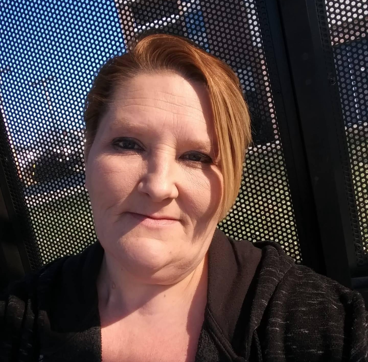 A woman is waiting at a bus stop, enjoying the warmth of the sun while looking at the camera.