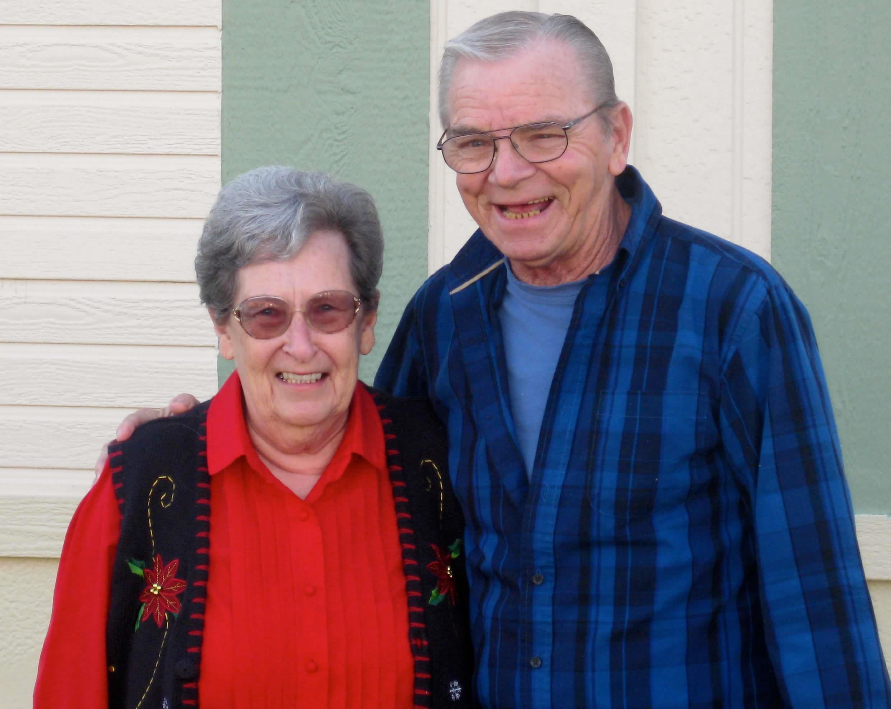 A joyful elderly couple stands closely, both smiling warmly at the camera in daylight.