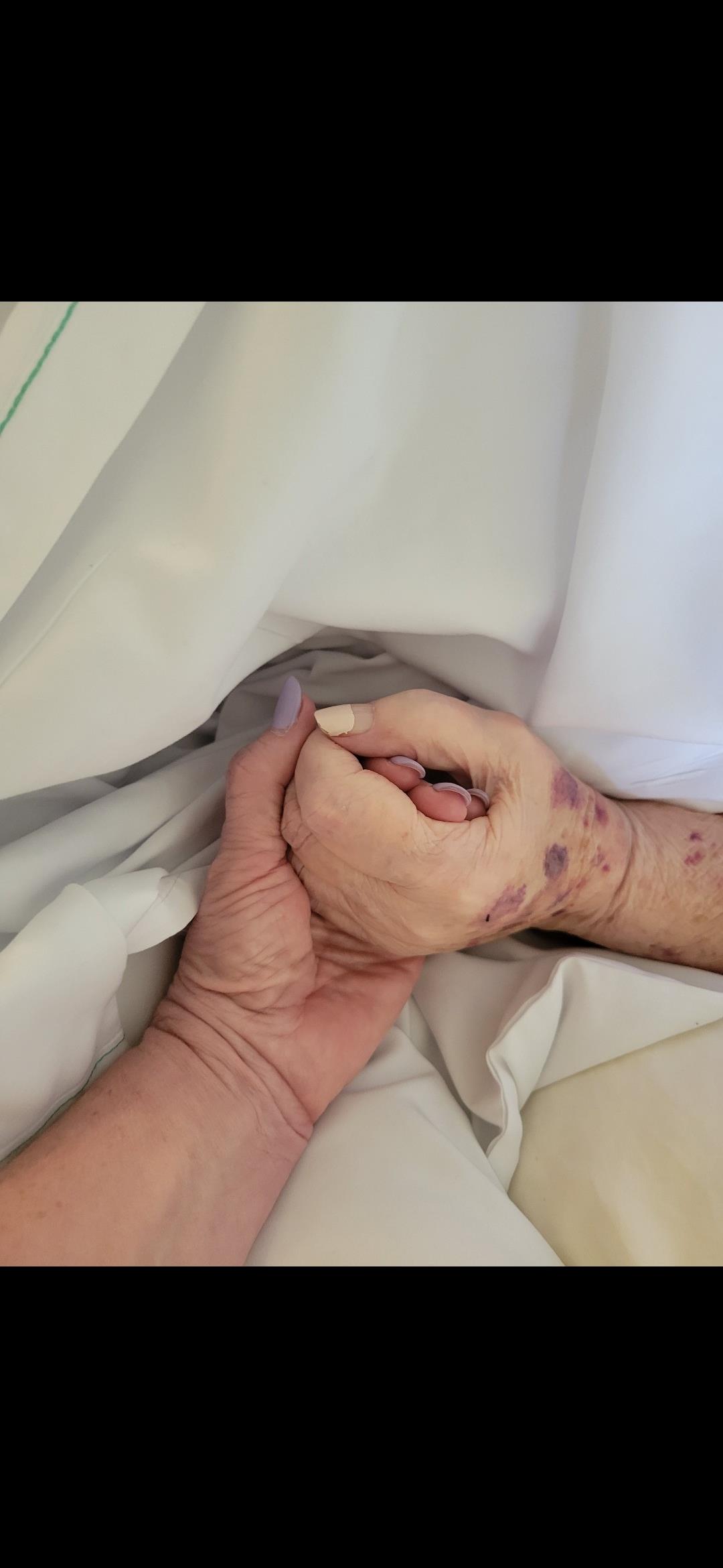A younger hand holds the hand of an elderly person in a hospital, showing tenderness and support.