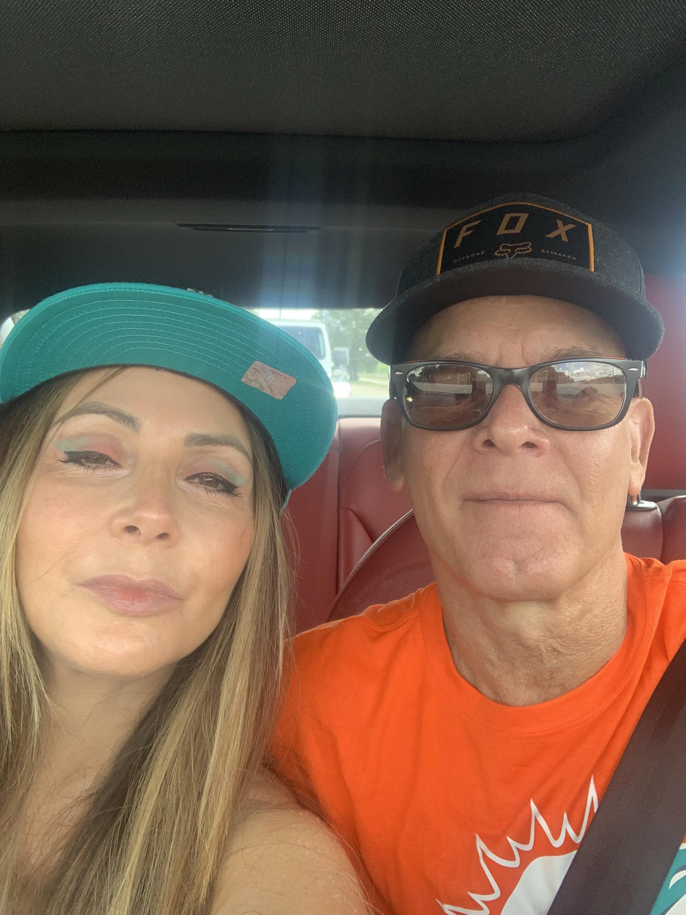 Two people take a selfie inside a car, both wearing vibrant caps and smiling joyfully.