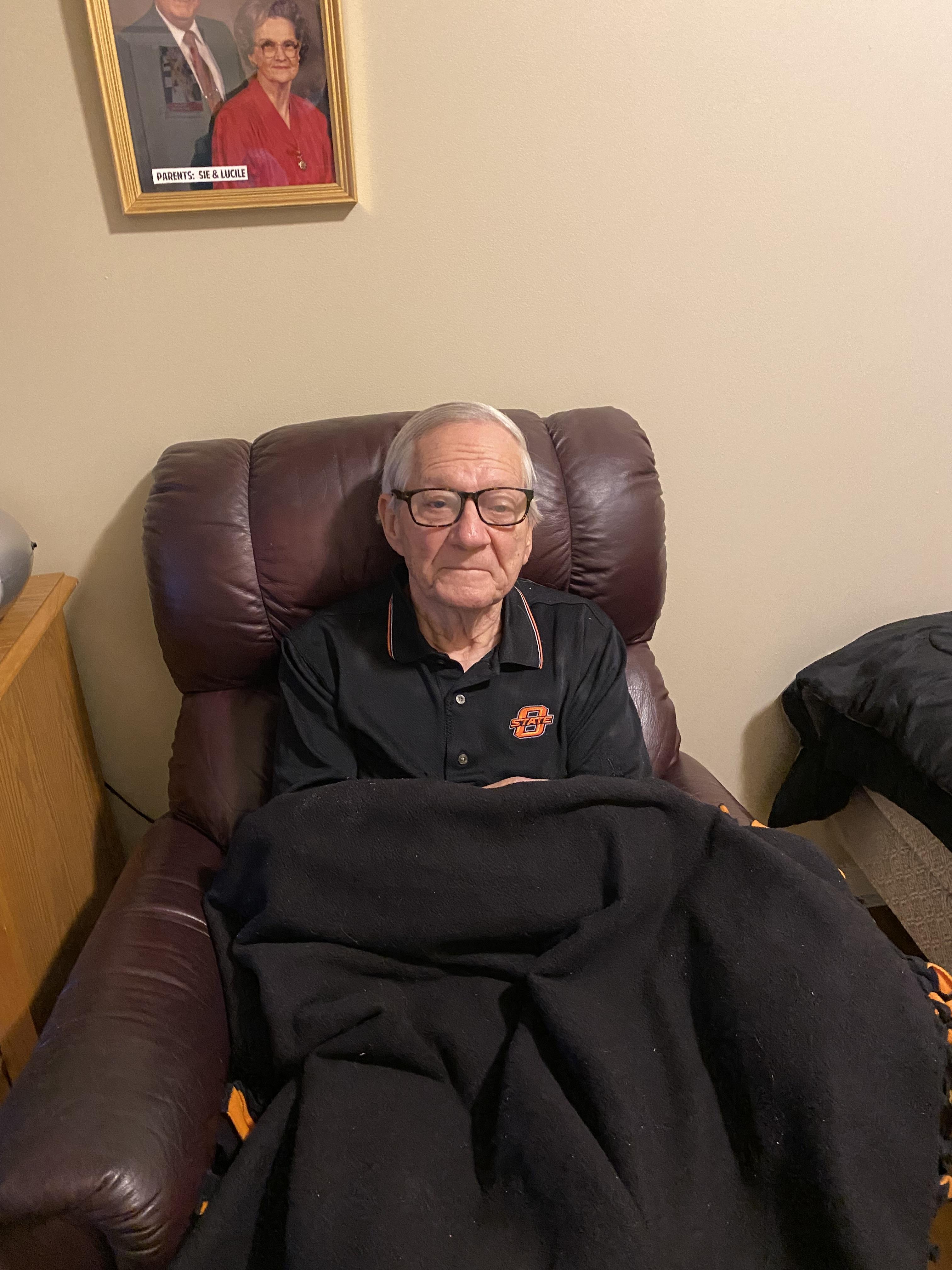 Older gentleman sits in a recliner wearing glasses, wrapped in a blanket, reflecting tranquility.