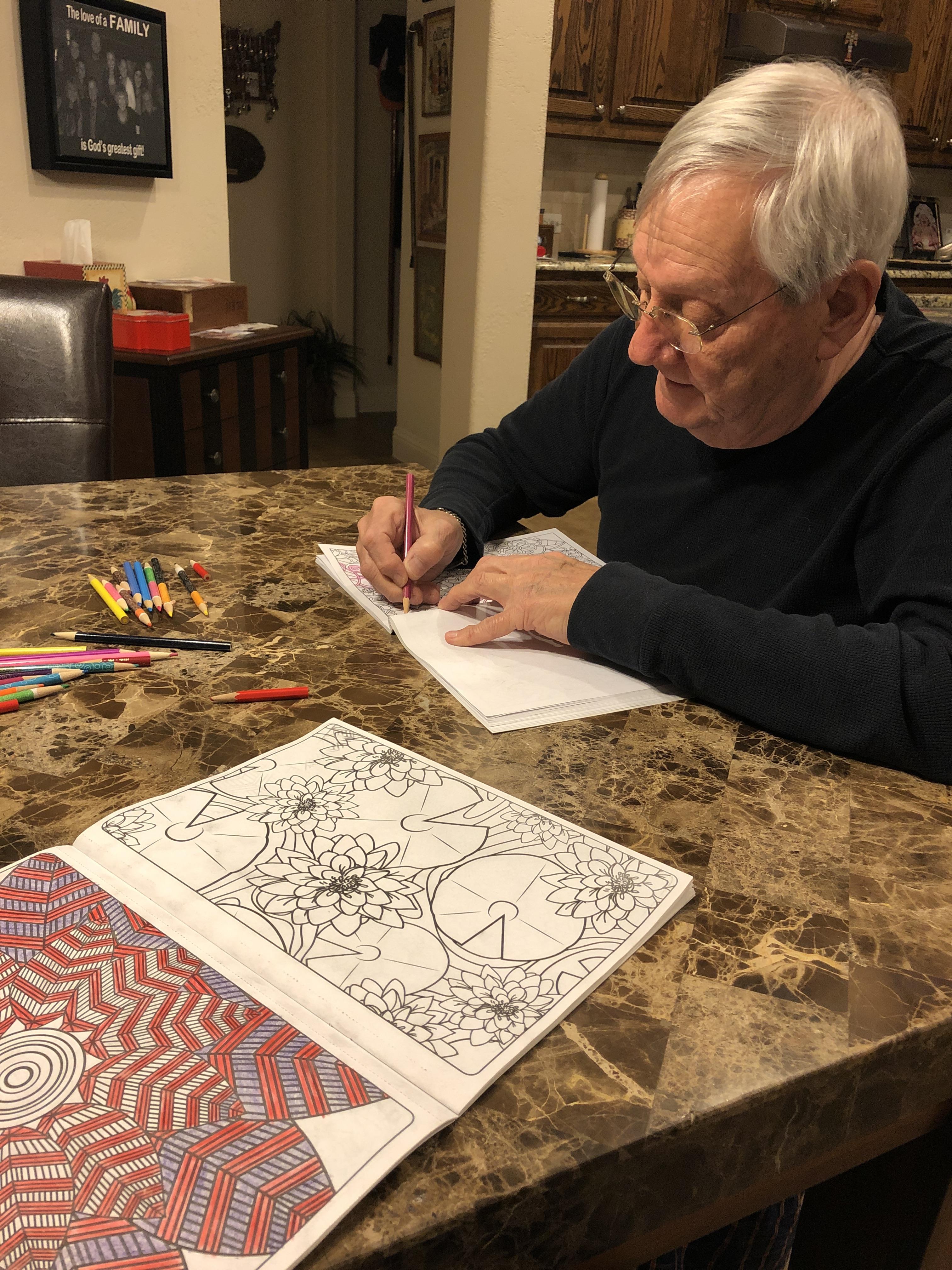 A man is concentrating on coloring intricate patterns at a table filled with art supplies.