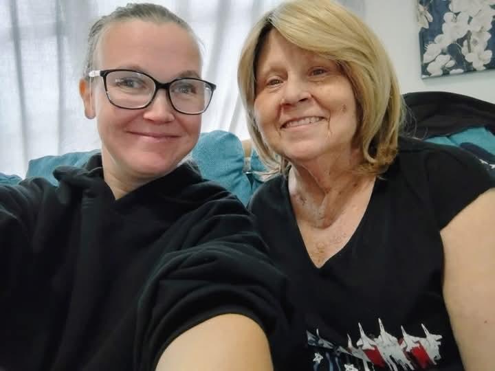 Two women share a joyful moment indoors, smiling at the camera in a cozy living room setting.