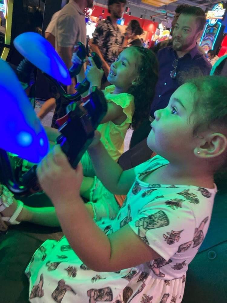 Two young girls play games at an arcade, fully engaged with their controllers and smiling.