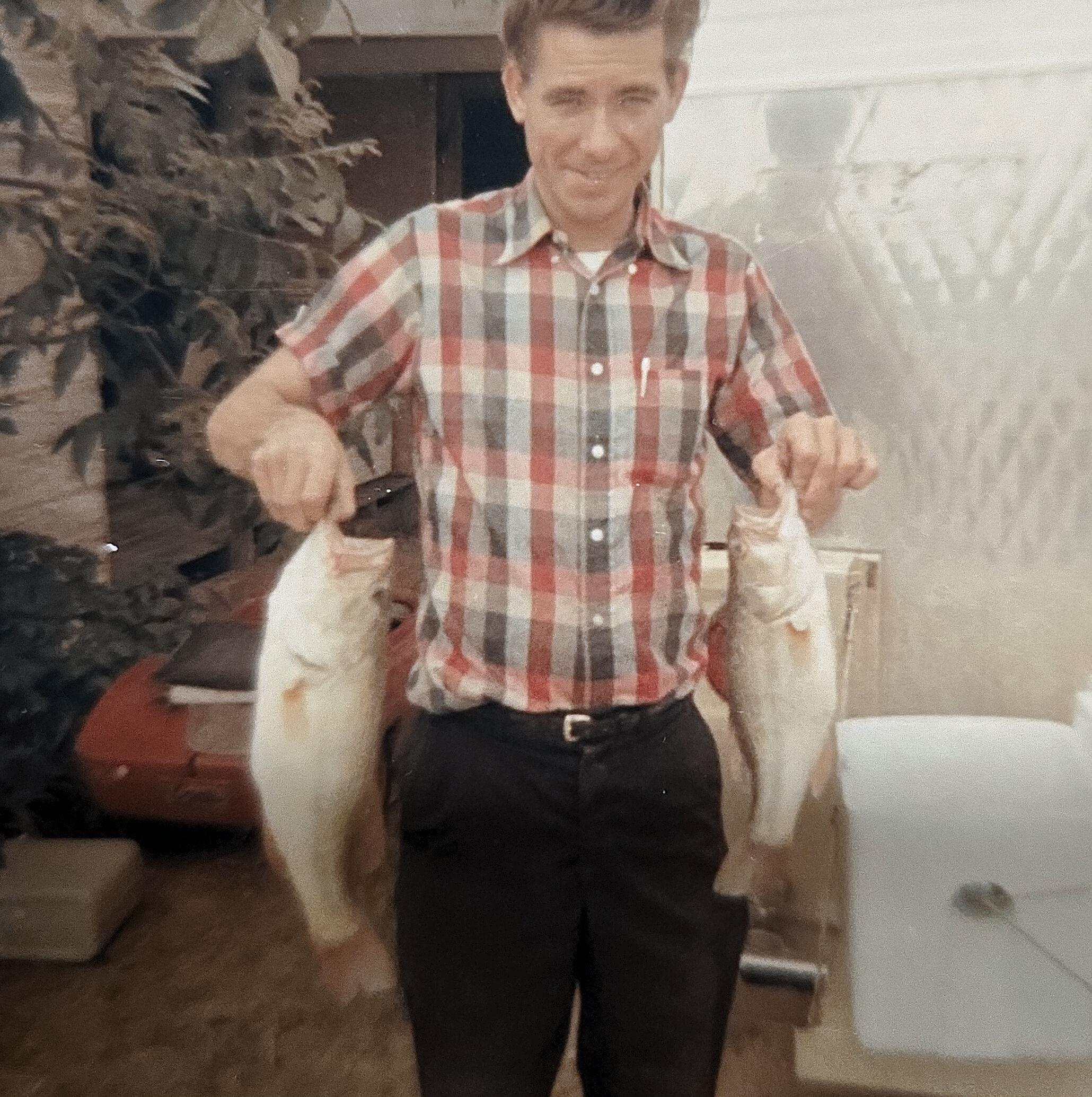A man holds two large fish in each hand, smiling against a natural backdrop and vintage car.