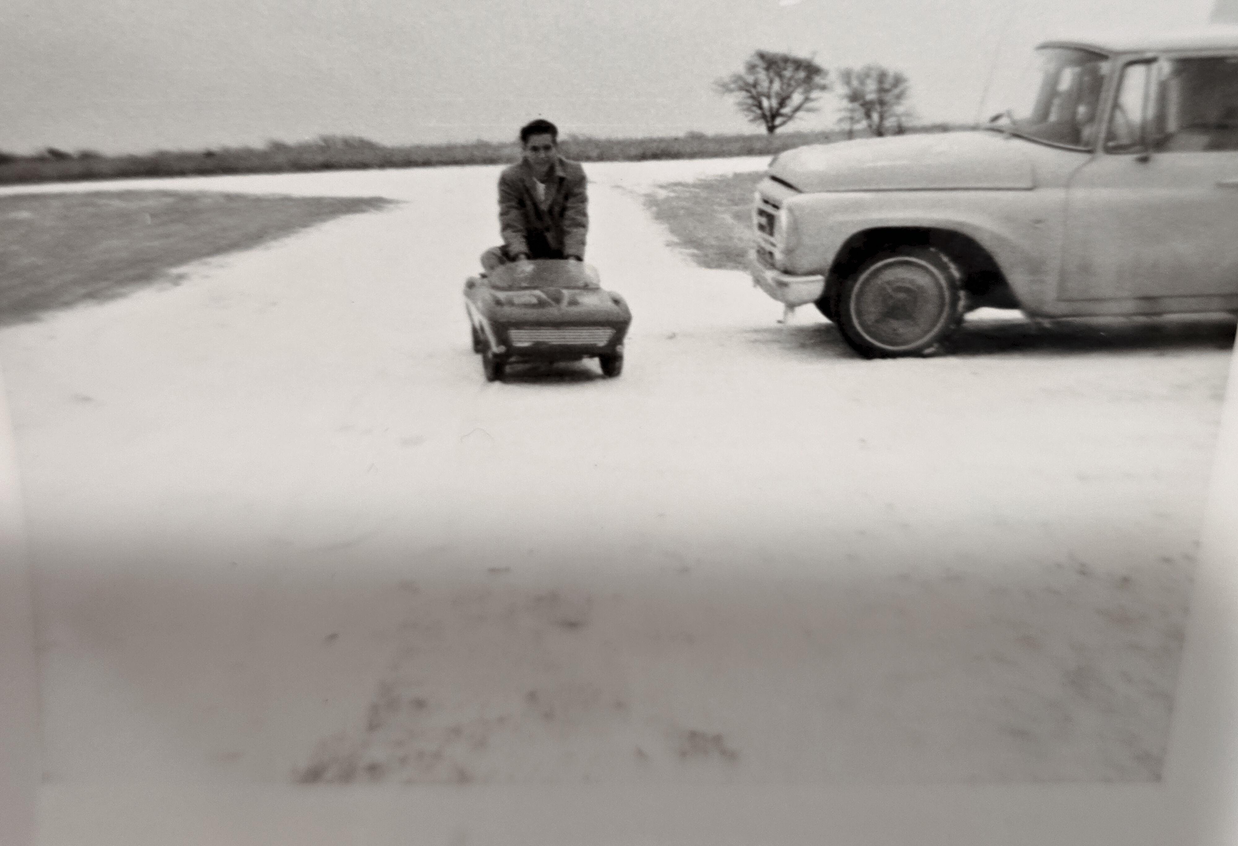 A boy is pulling a small toy car through snow on a rural road beside an old truck on a winter day.