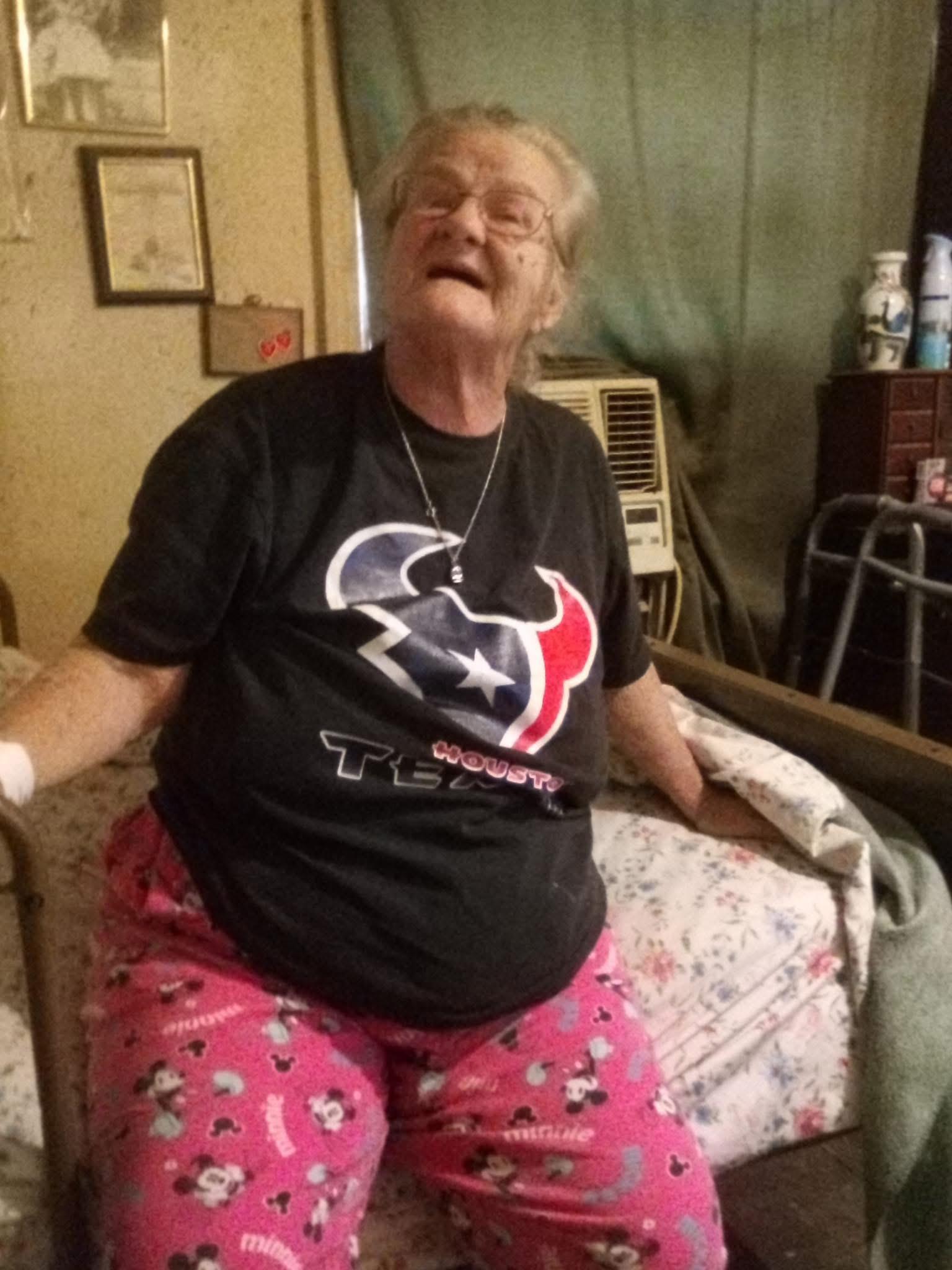 An elderly woman sits on her bed, smiling and wearing cozy pajamas while enjoying her day at home.
