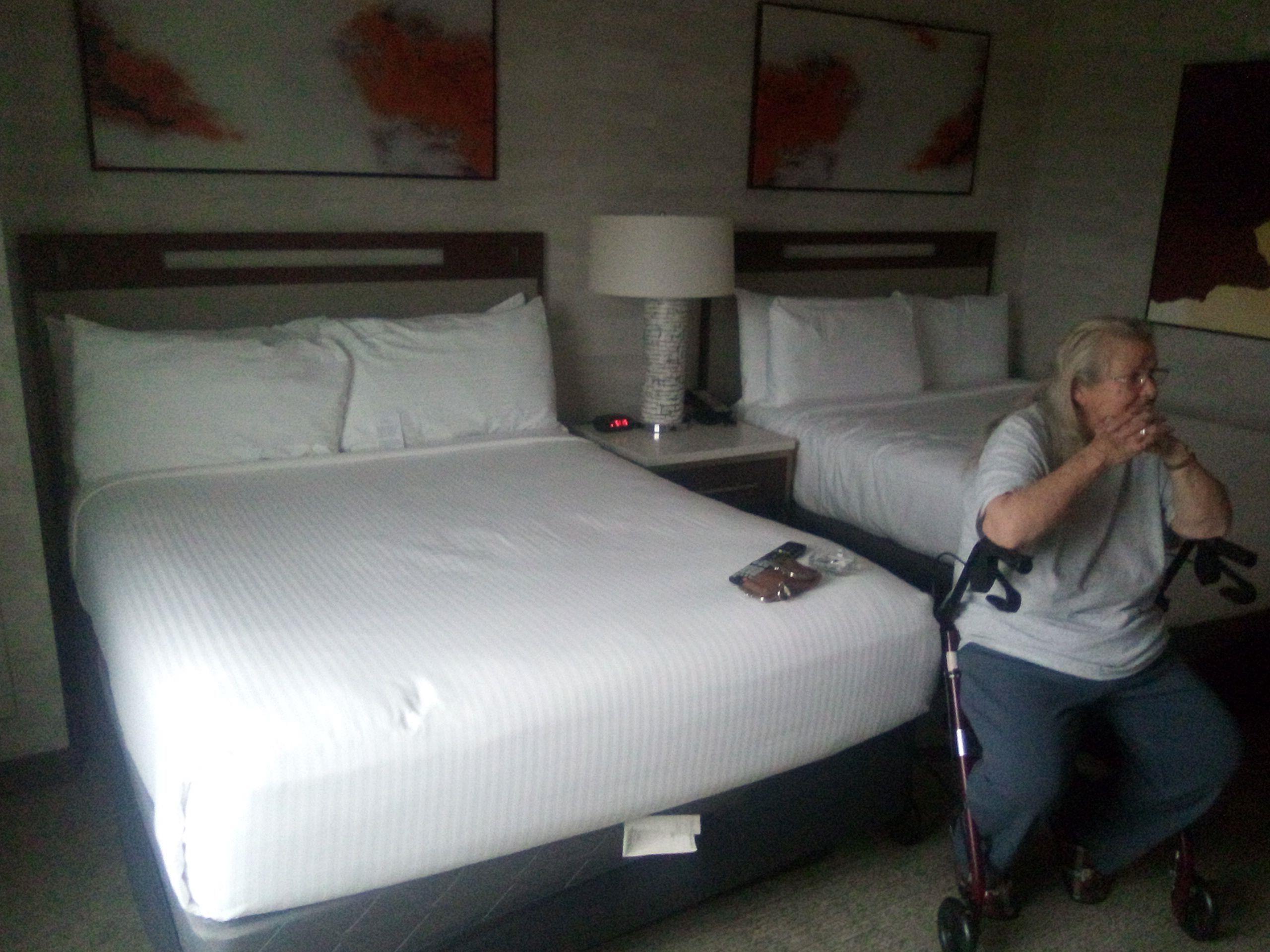 An elderly woman sits on a walker beside two neatly made beds in a hotel room.